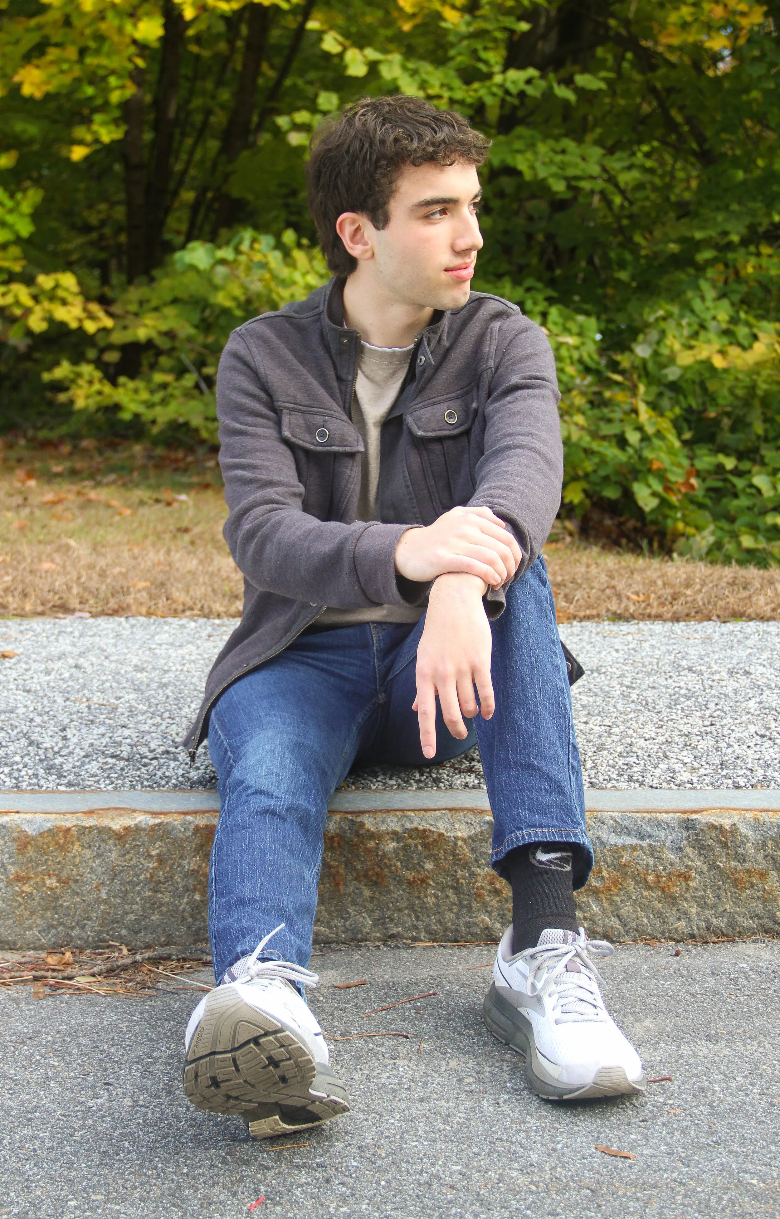 A young man sitting on a curb in an outdoor park, wearing a gray jacket, jeans, and sneakers, with trees and foliage in the background.