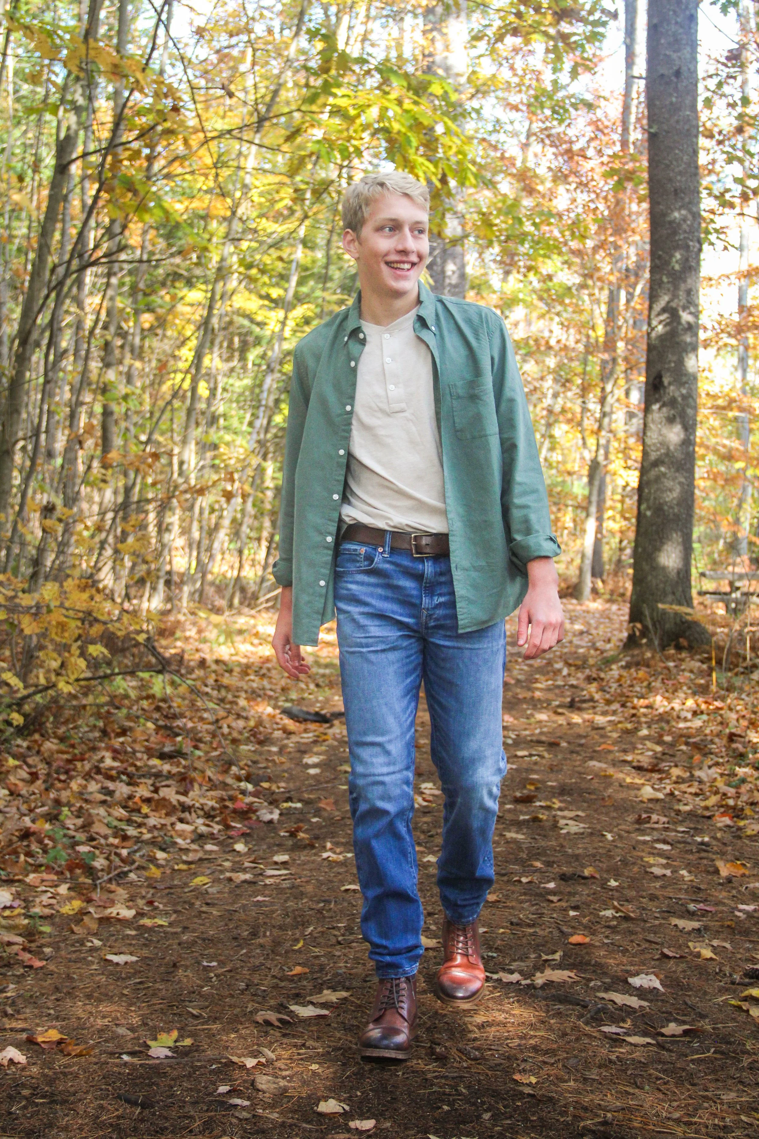 A young man walking on a leaf-covered trail in a forest during autumn, smiling and enjoying nature.