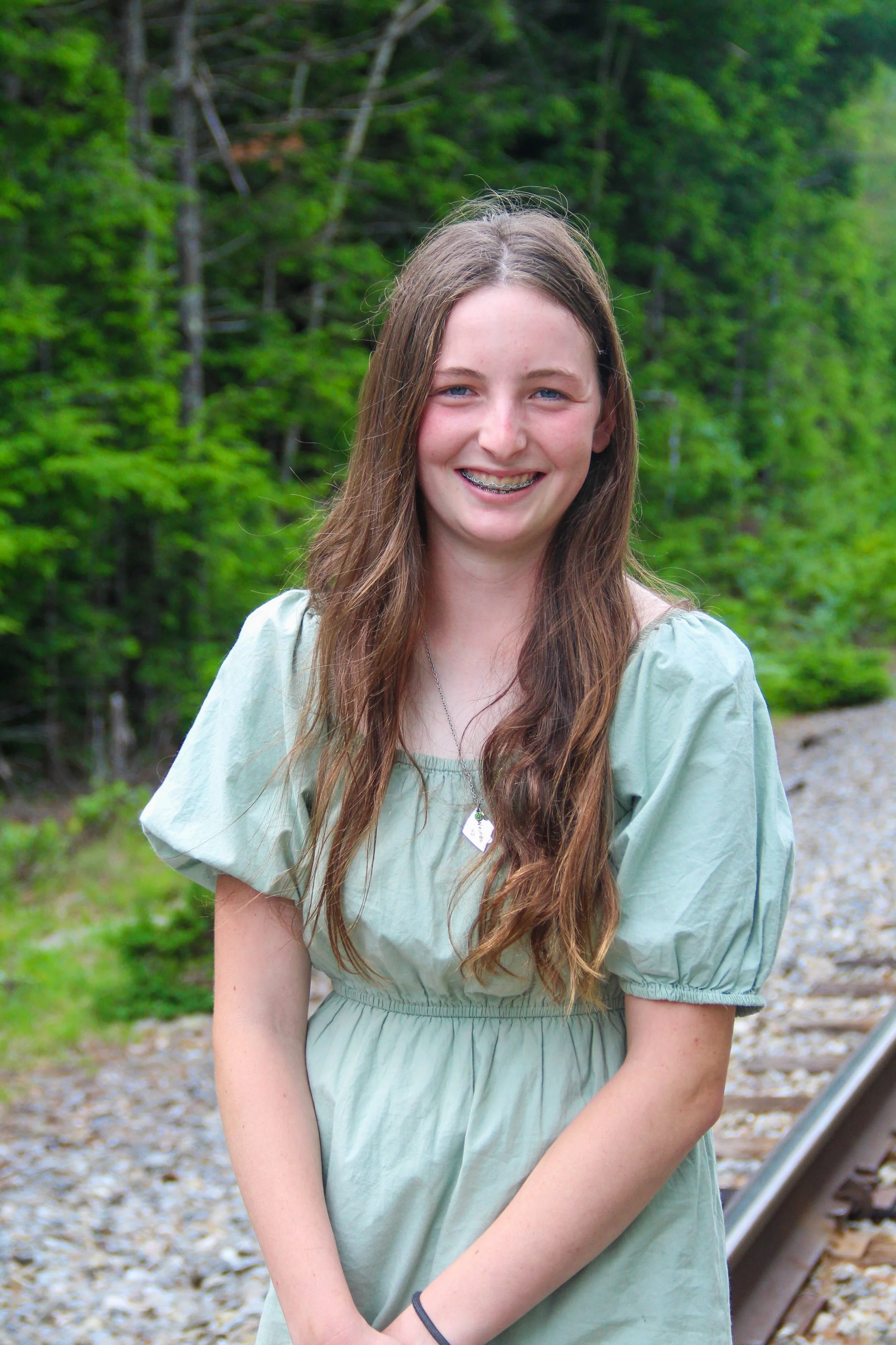 A girl standing outdoors near a railway track, smiling at the camera with long brown hair, wearing a light green dress and a necklace, with green trees in the background.
