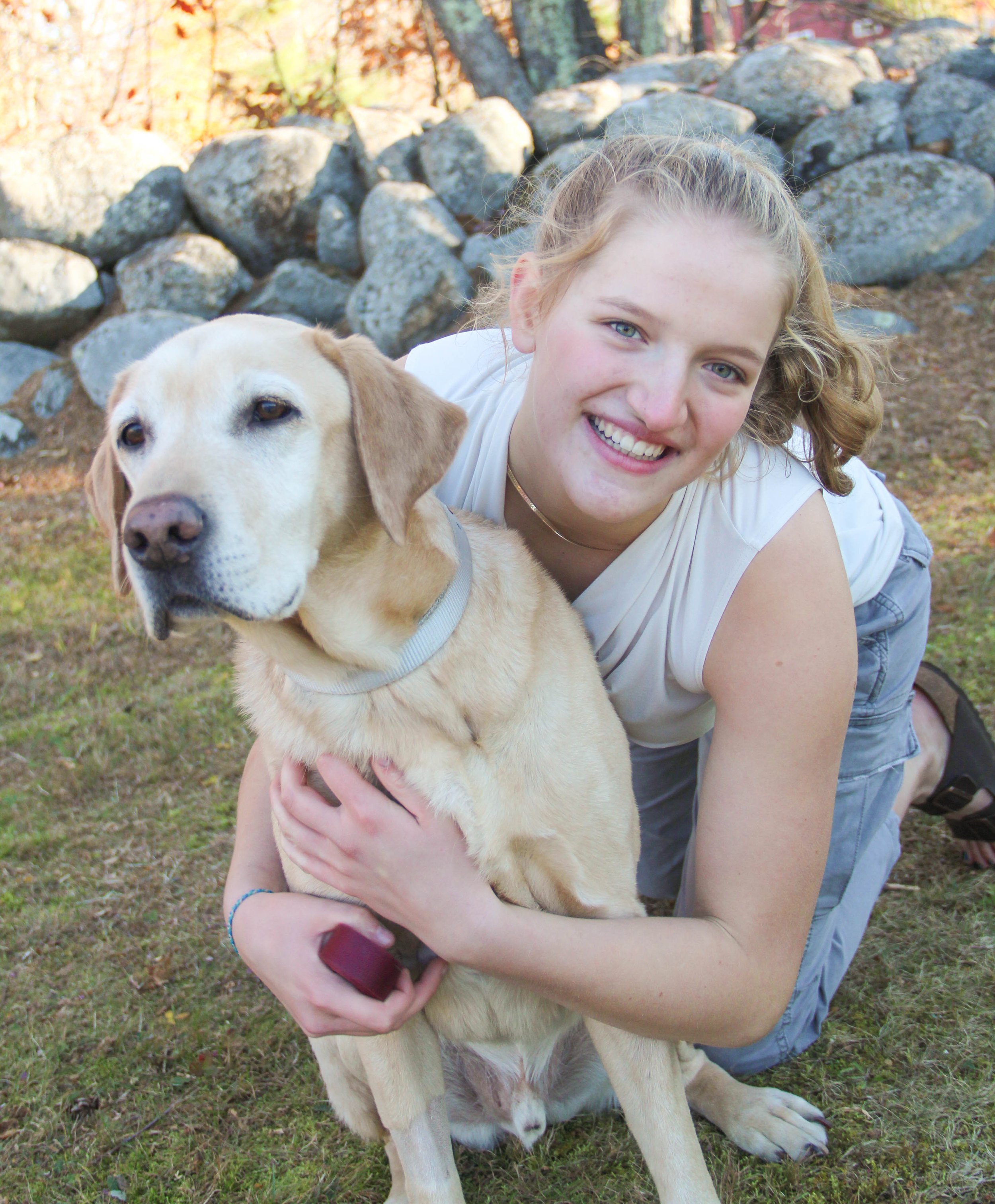 A young woman with blonde curly hair smiling and hugging a yellow Labrador retriever outside on grass with rocks and trees in the background.