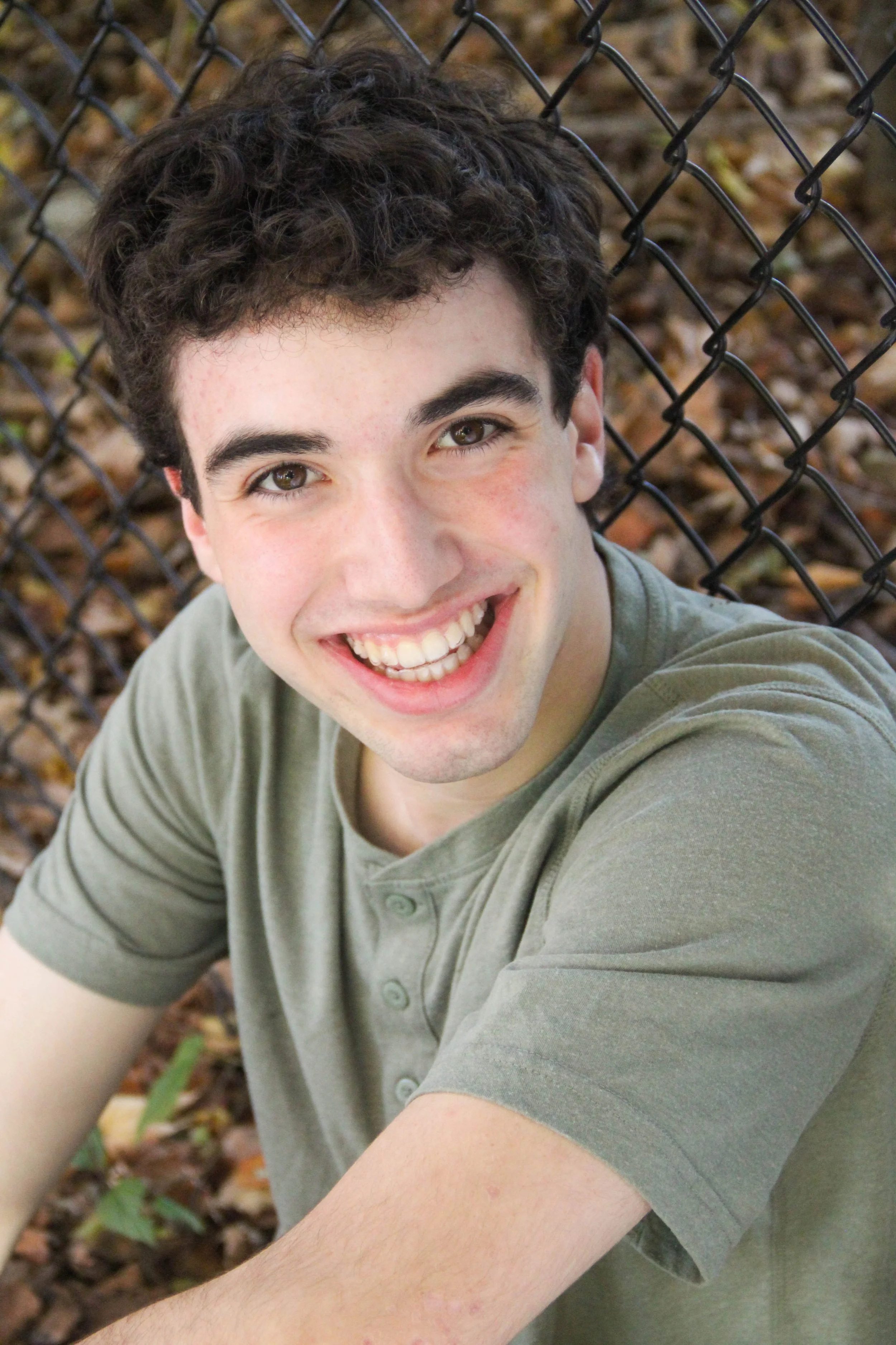Smiling young man with curly dark hair, wearing a green t-shirt, sitting outdoors with fallen leaves and a chain-link fence in the background.