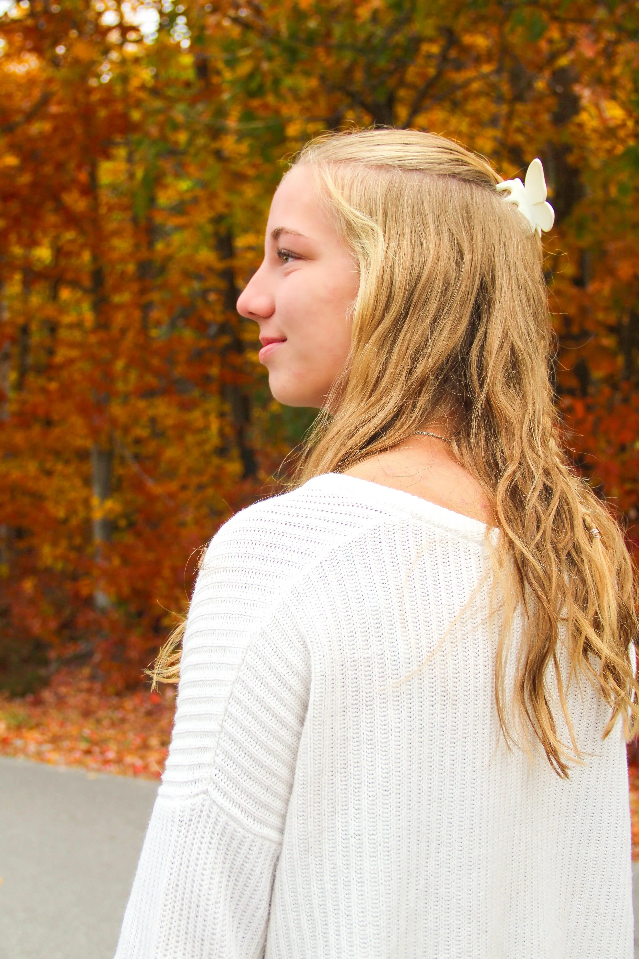 Profile of a young woman with blonde hair wearing a white sweater, standing outdoors with autumn foliage in the background.