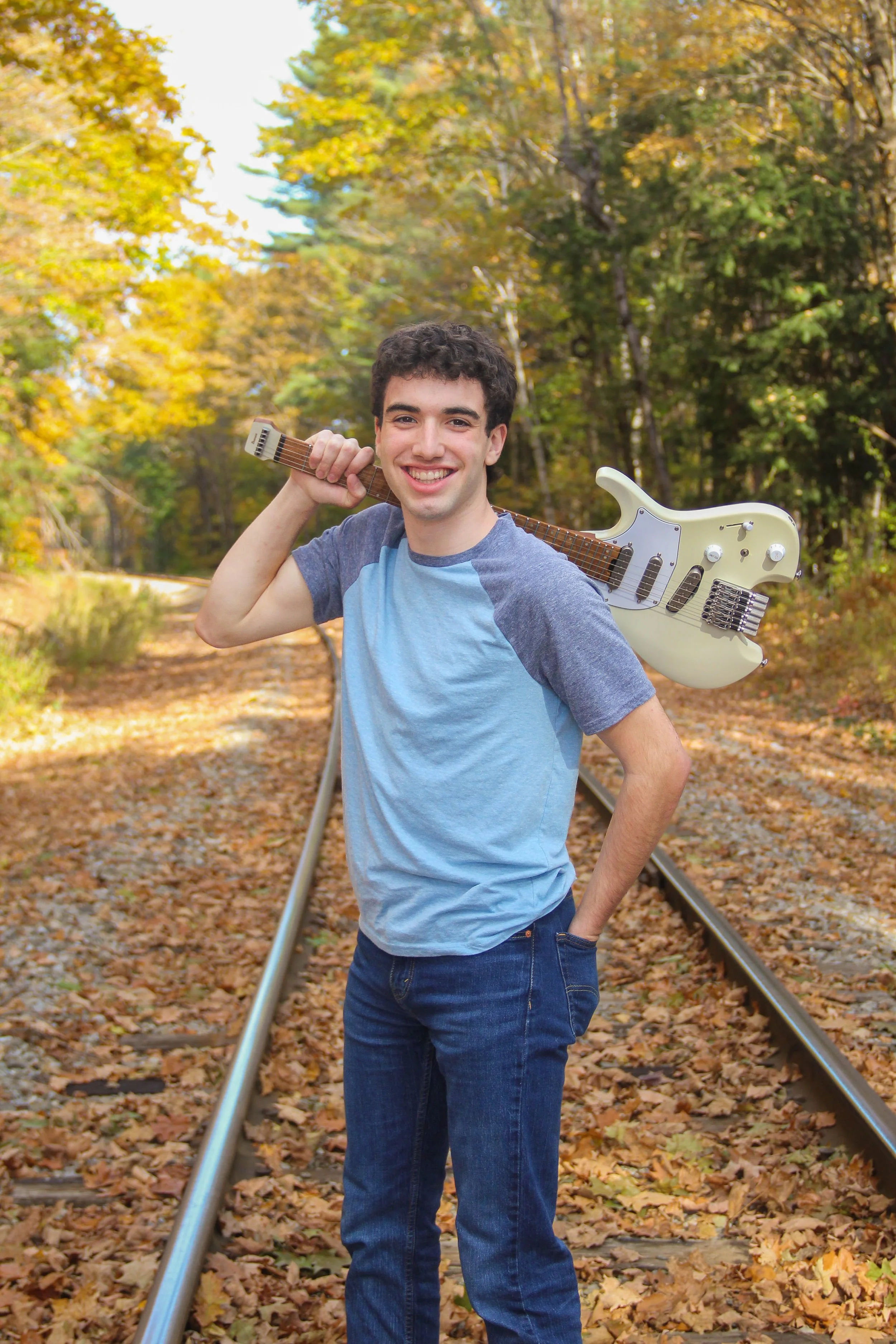 A young man with dark curly hair and a bright smile, wearing a blue and gray T-shirt and blue jeans, stands on a set of train tracks surrounded by autumn trees with colorful leaves. He has a guitar slung over his shoulder.