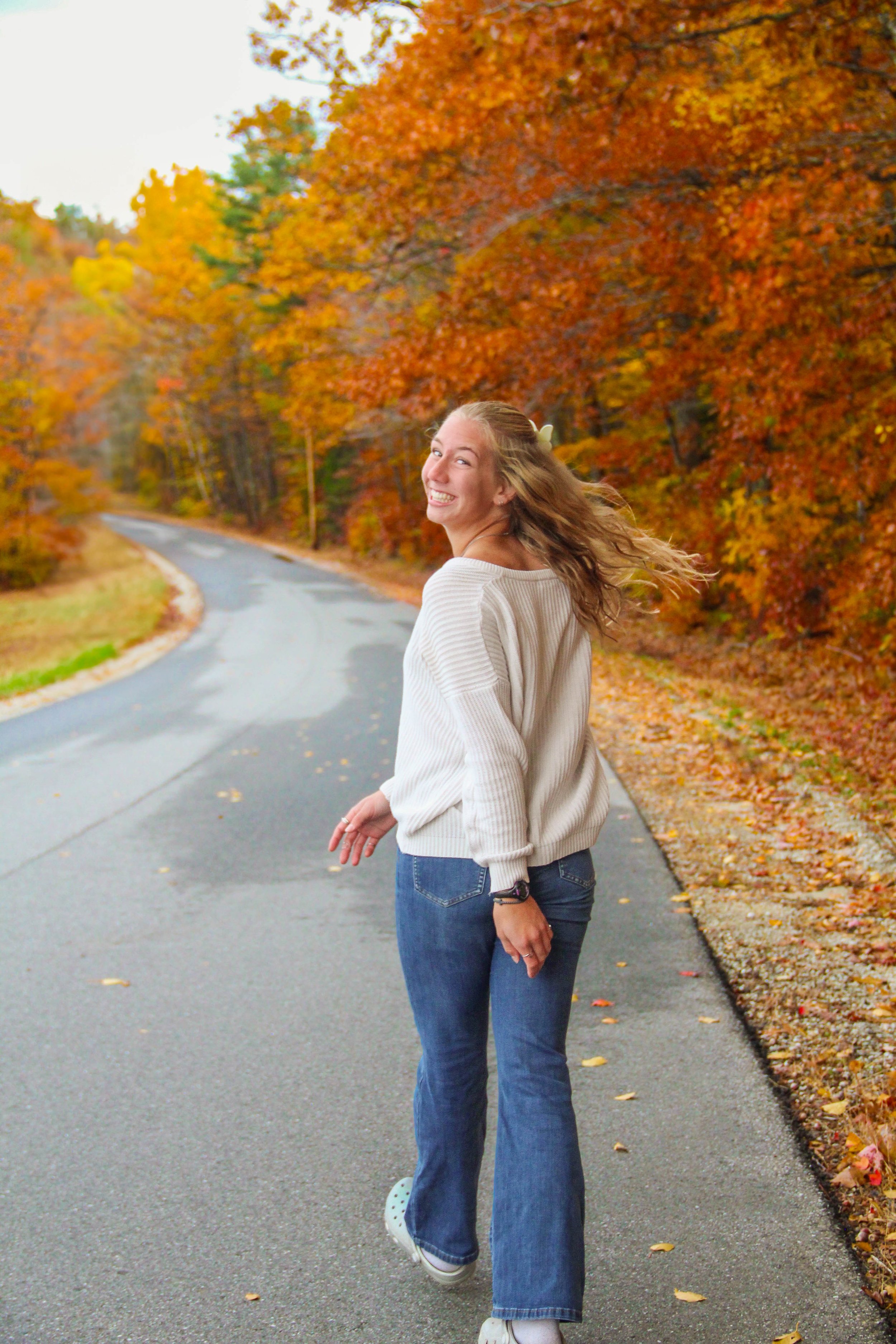 A young woman with long hair, wearing a white sweater, jeans, and sneakers, smiling and walking on an autumnal road lined with colorful fall trees.