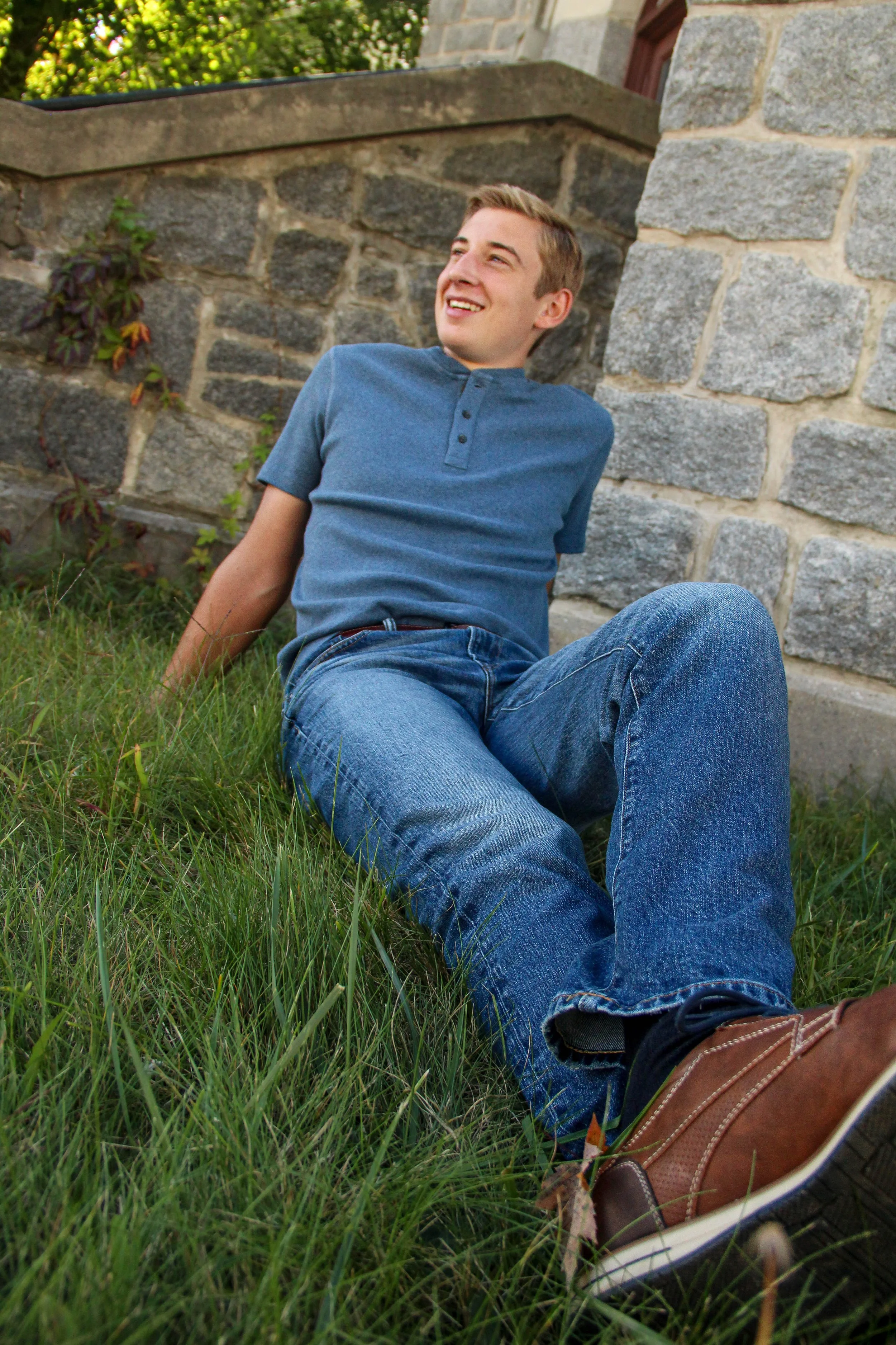 A young man wearing a blue shirt and jeans sitting on the grass, leaning against a stone wall and smiling.
