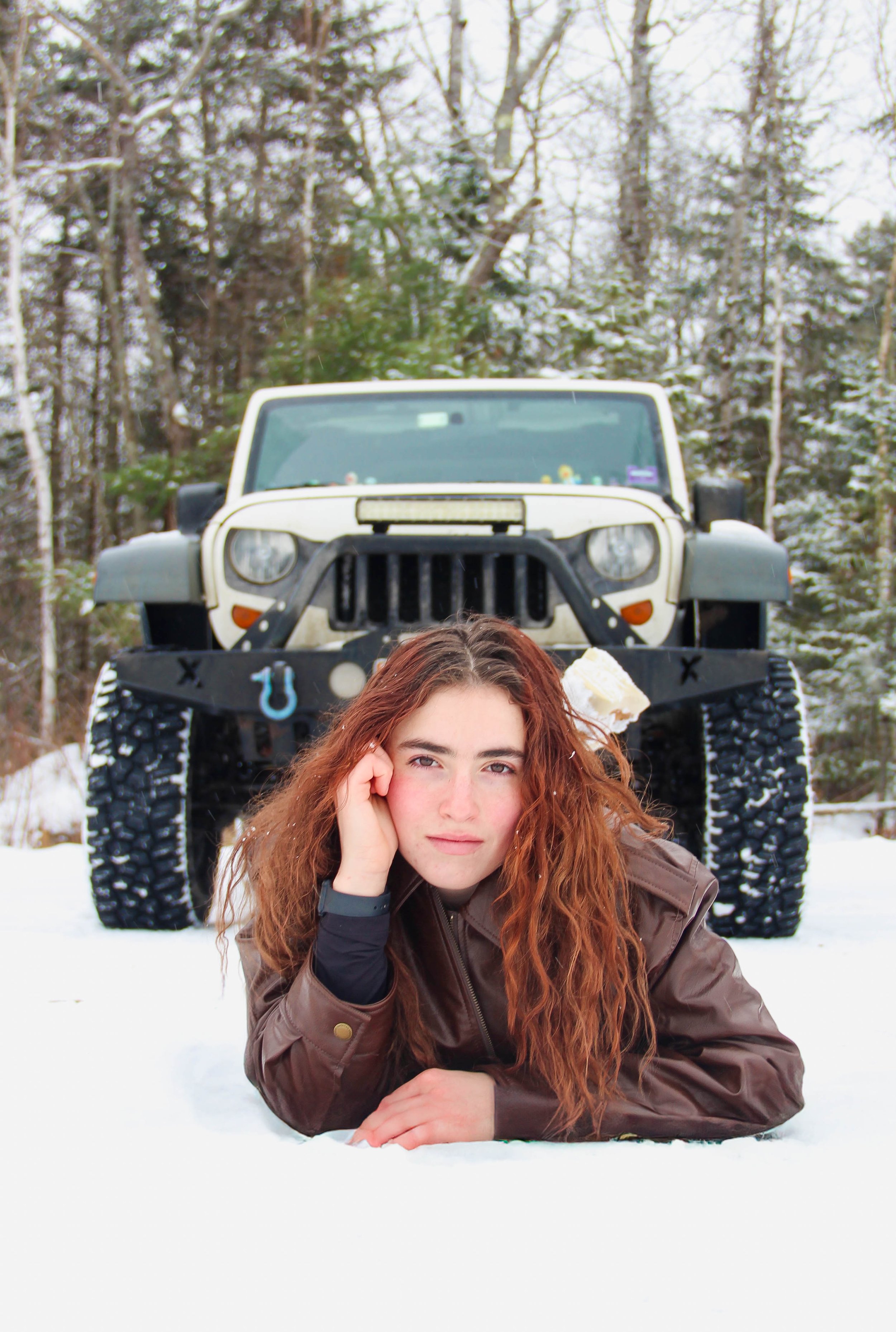 A young woman with long curly red hair lying on snow in front of a beige Jeep in a snowy forest.