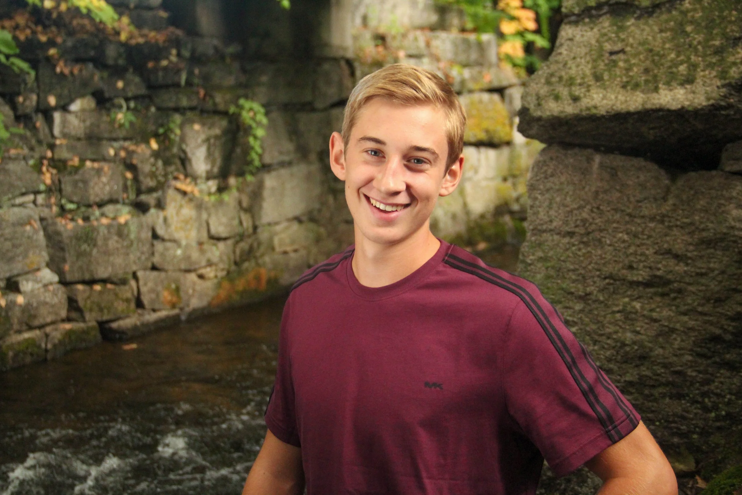 A smiling young man with blonde hair wearing a maroon t-shirt with black stripes on the shoulders, standing outdoors near a stone wall and a small stream.