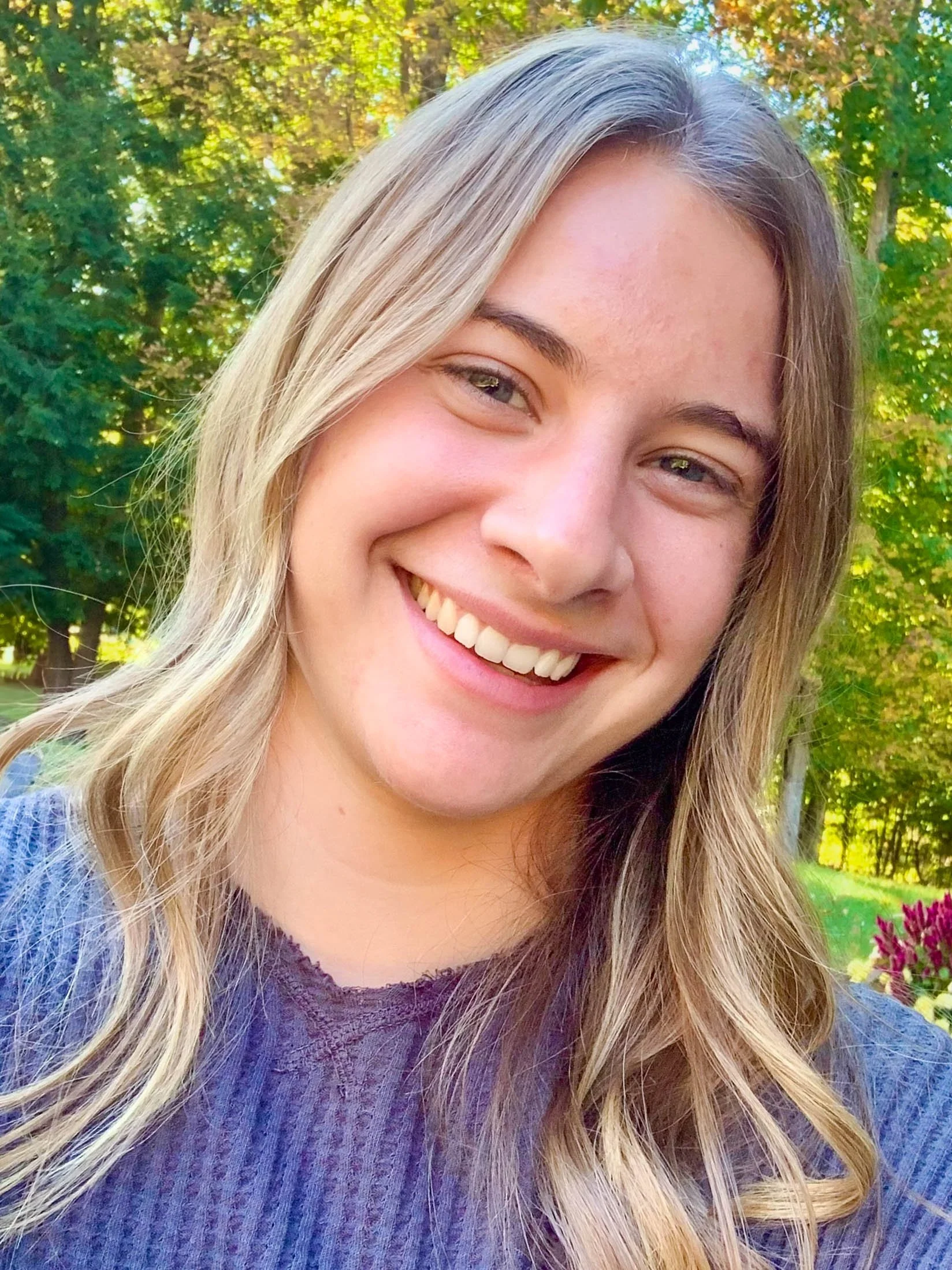 Smiling young woman with long blond hair outdoors in a park with trees and flowers in the background.
