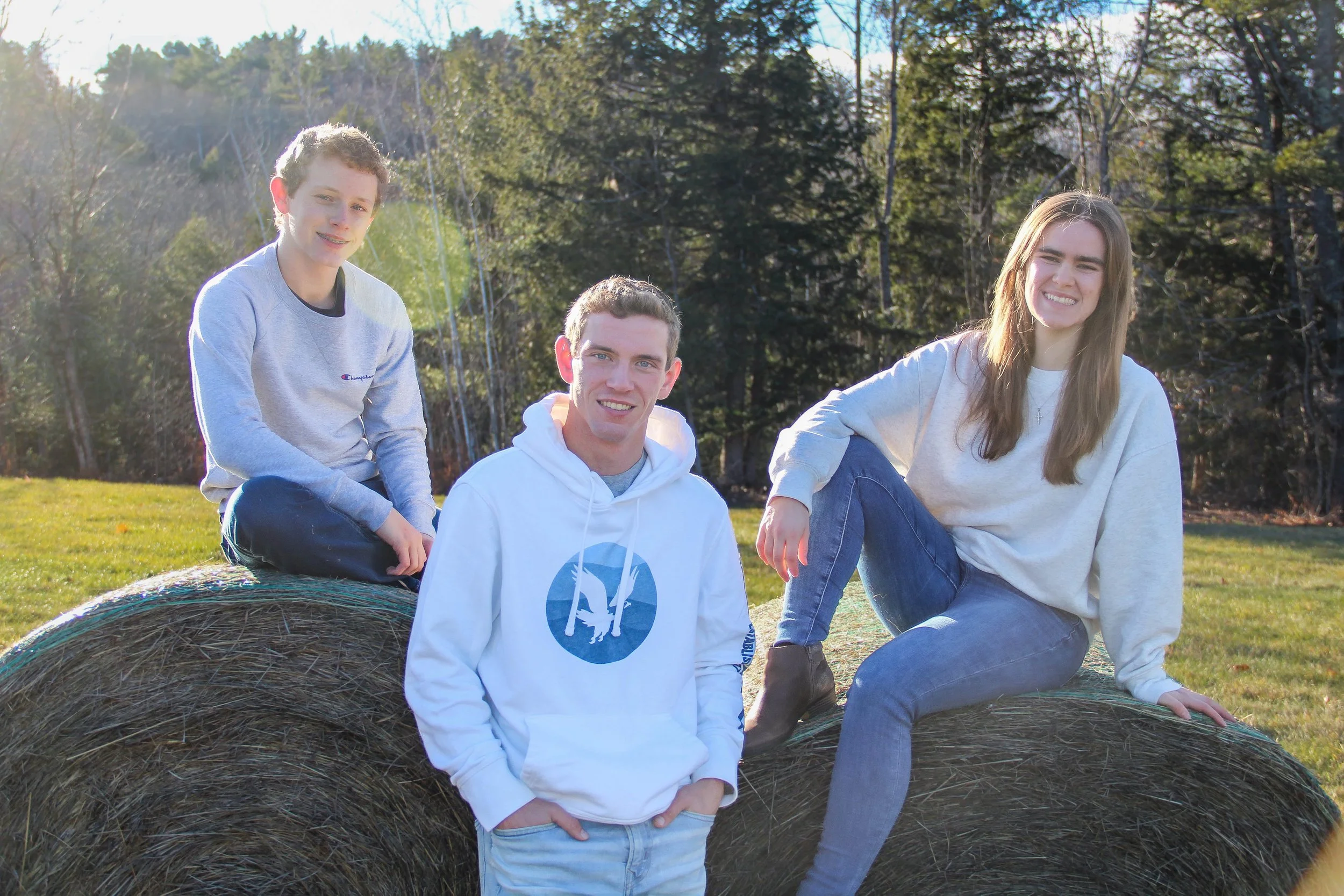 Three teenagers sitting and leaning on a large hay bale outdoors in a field with trees in the background.