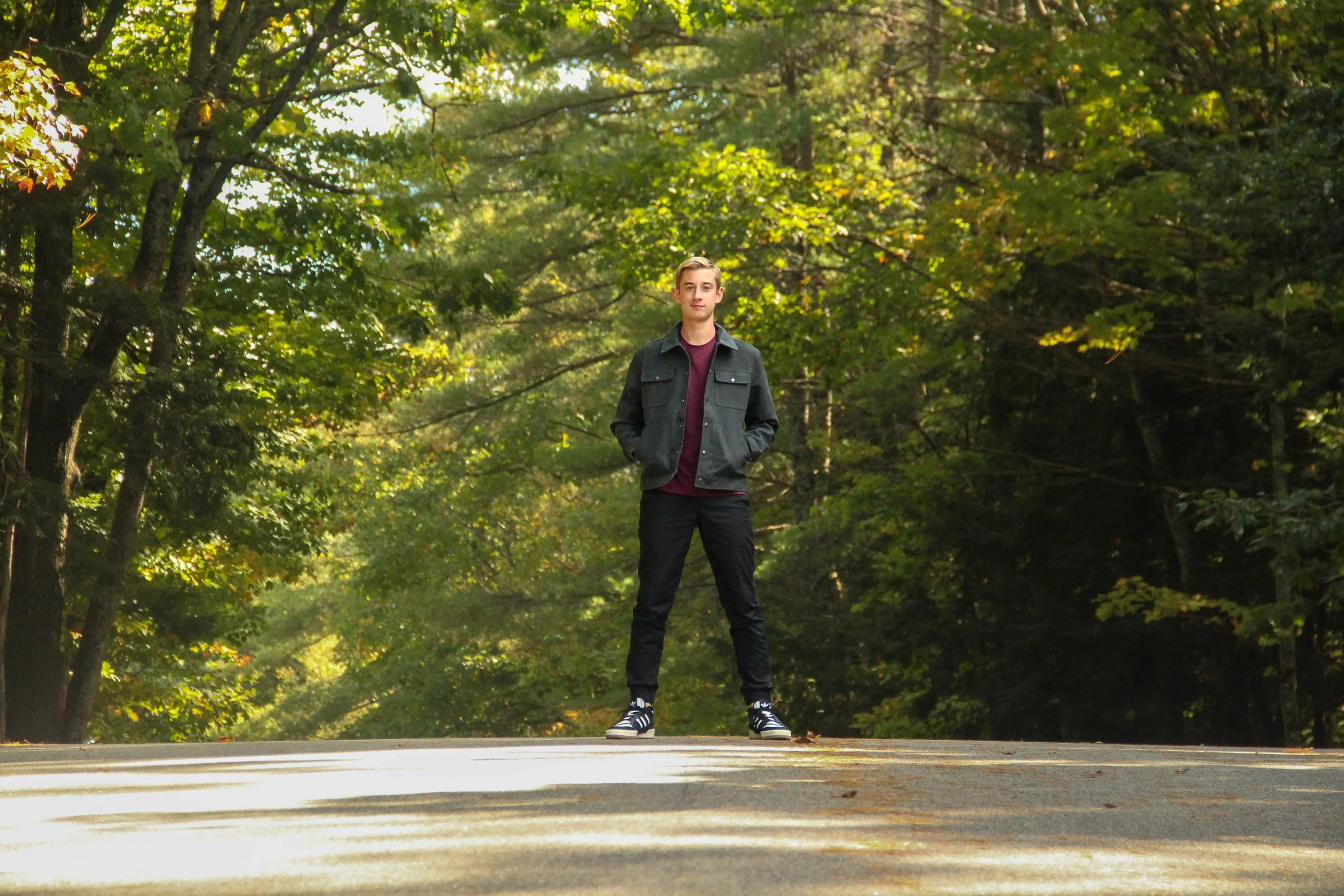 A young man standing in the middle of a wooded road with green trees and sunlight in the background.