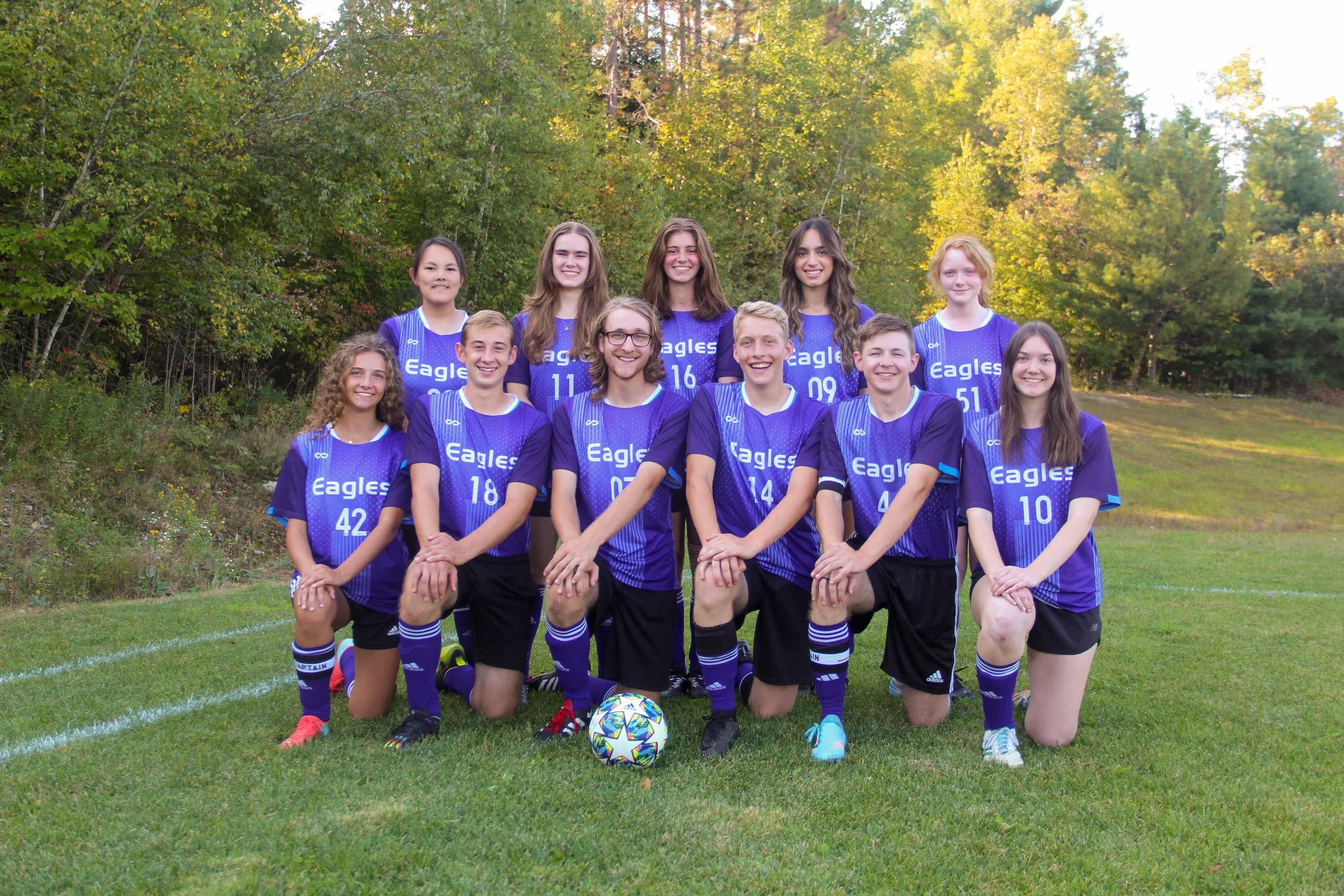 A group of eleven young girls and boys in purple soccer jerseys kneeling and standing on a soccer field with a colorful soccer ball in front, surrounded by trees with autumn foliage.
