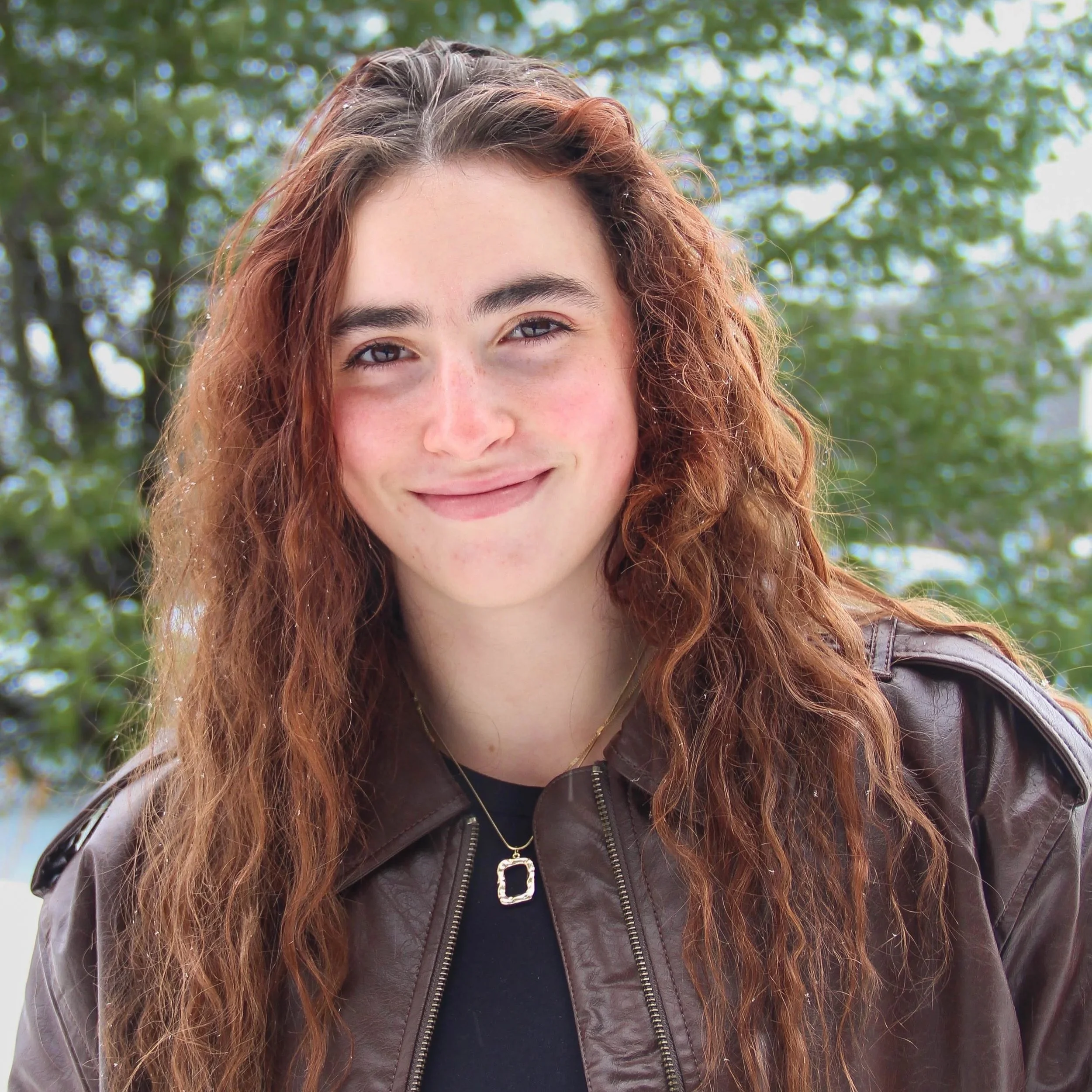 A young woman with long, curly red hair, smiling in an outdoor setting with green trees in the background.