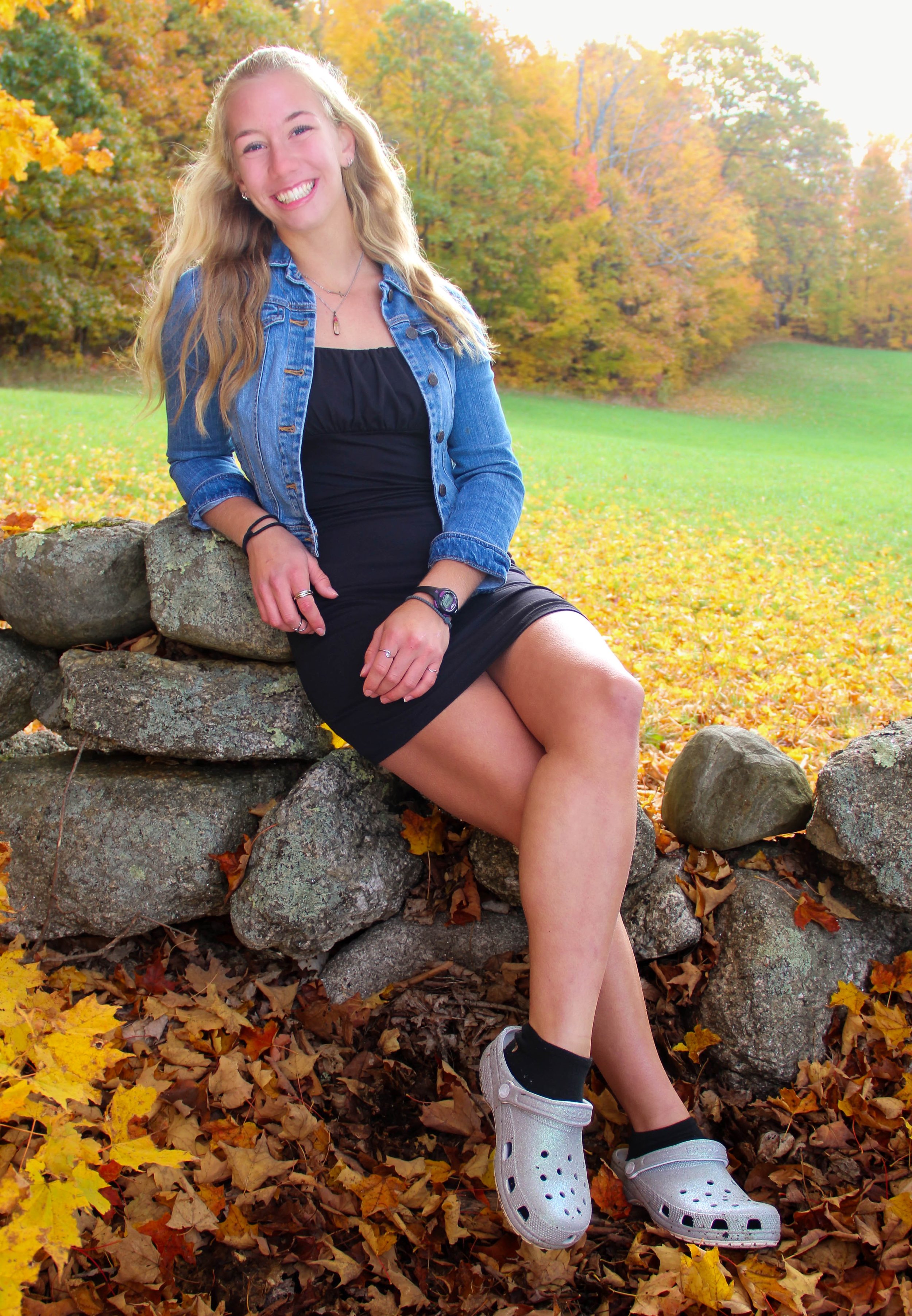 A young woman with long blonde hair sitting on a stone wall in a park during fall season, smiling and wearing a black dress, denim jacket, and white Crocs, with fallen leaves on the ground and autumn-colored trees in the background.