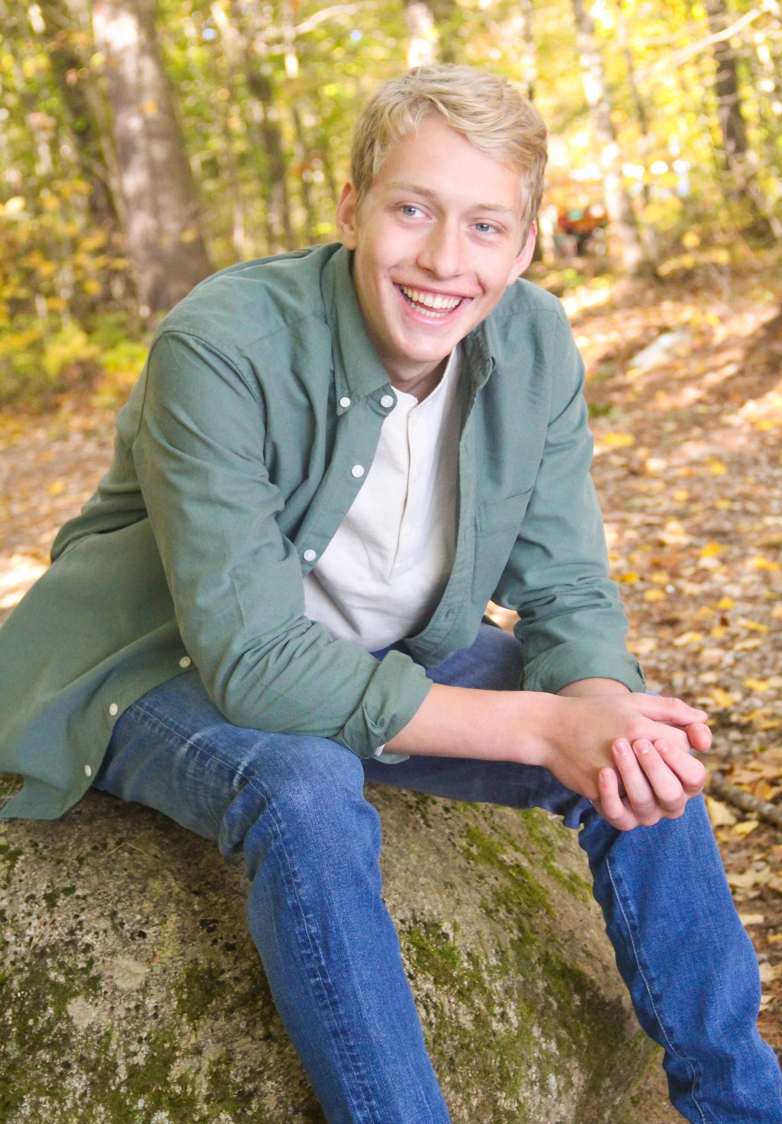 A young man with blonde hair smiling, sitting on a moss-covered rock in a forest with autumn leaves.
