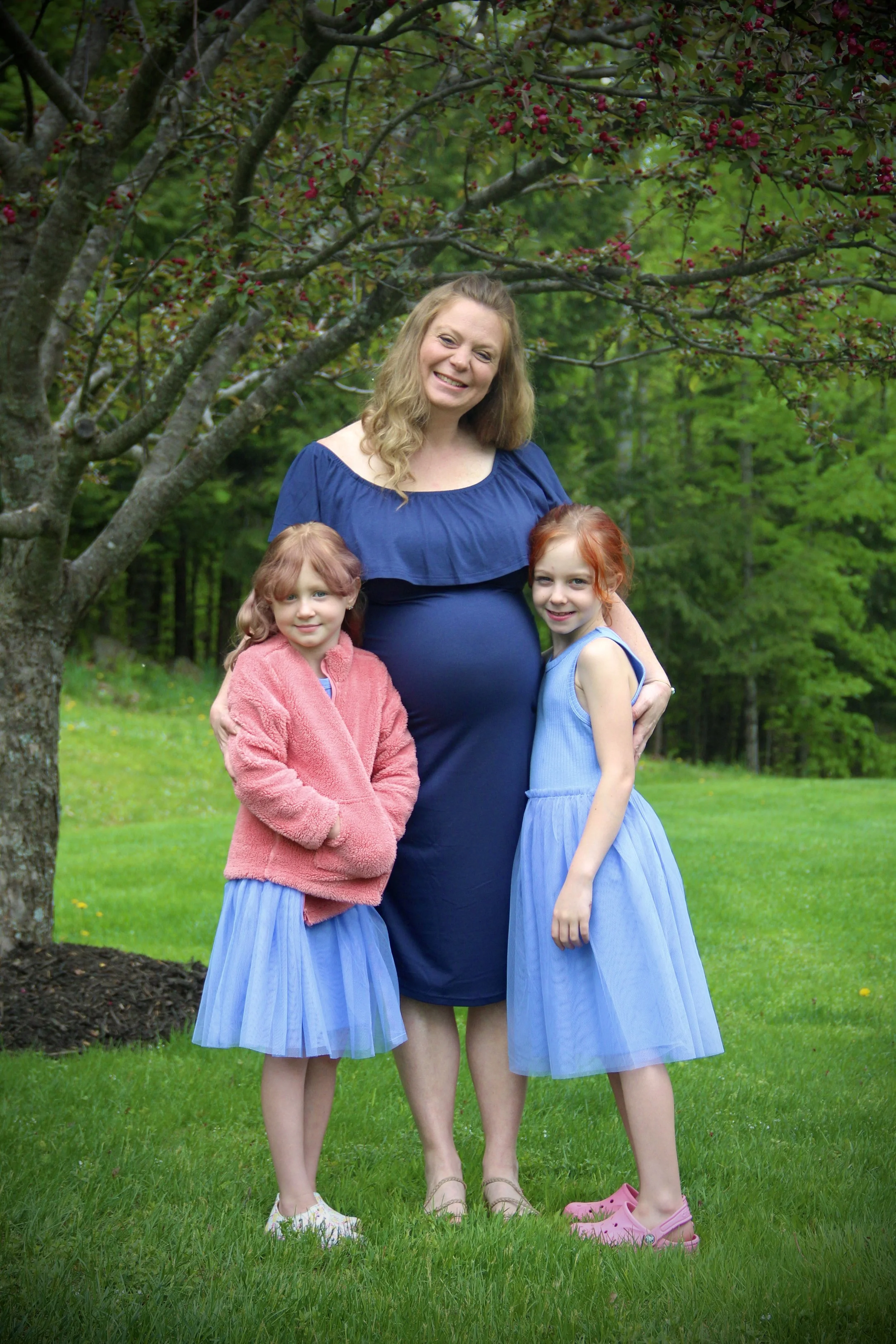 A woman and two young girls standing outdoors on green grass under a tree with red berries. The woman is smiling and wearing a blue dress. The girls are dressed in blue skirts; one in a pink jacket, the other in a blue dress, and pink shoes.
