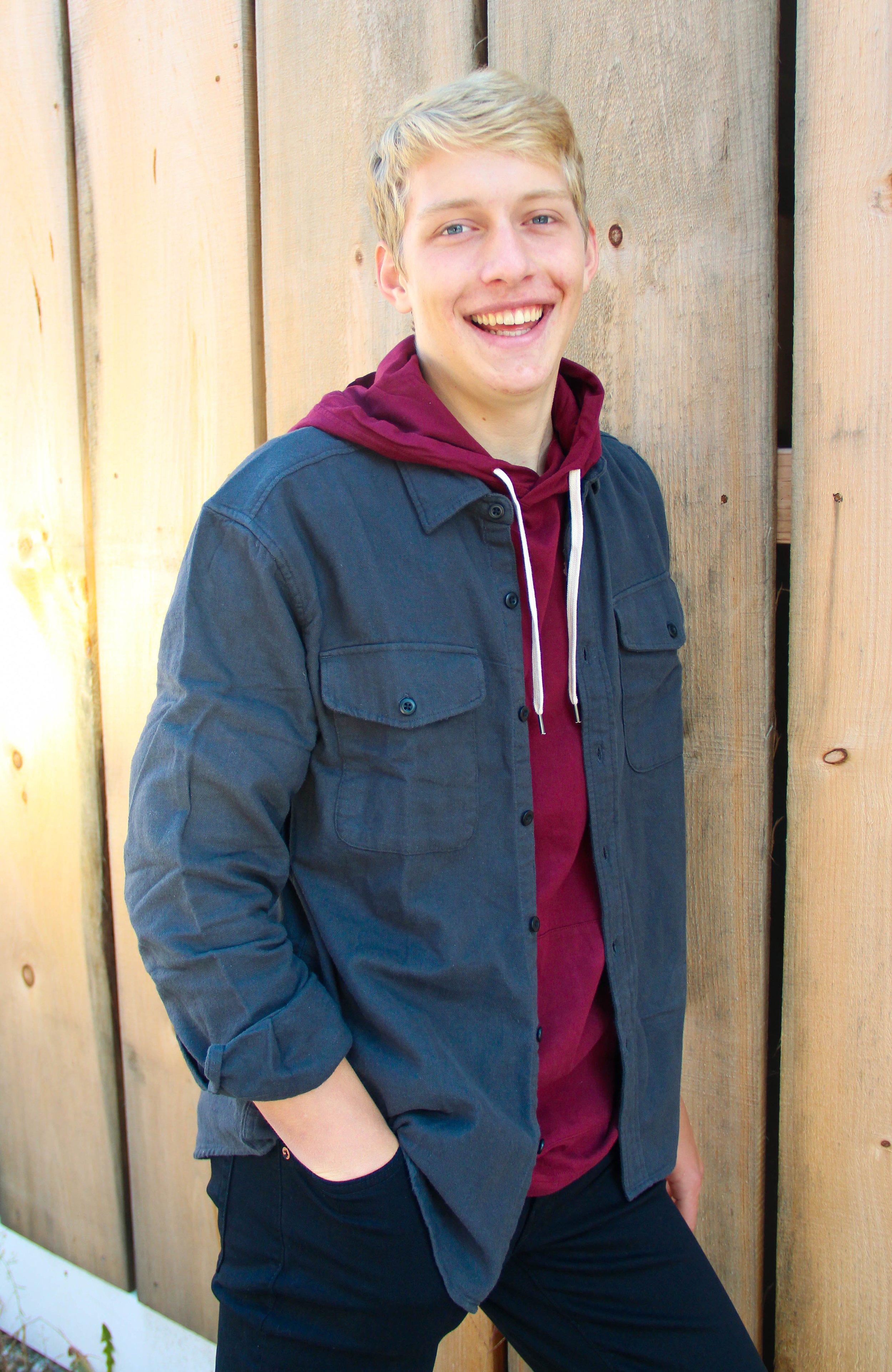 A young man with blond hair and a big smile, wearing a dark jacket over a maroon hoodie, standing outdoors against a wooden fence.