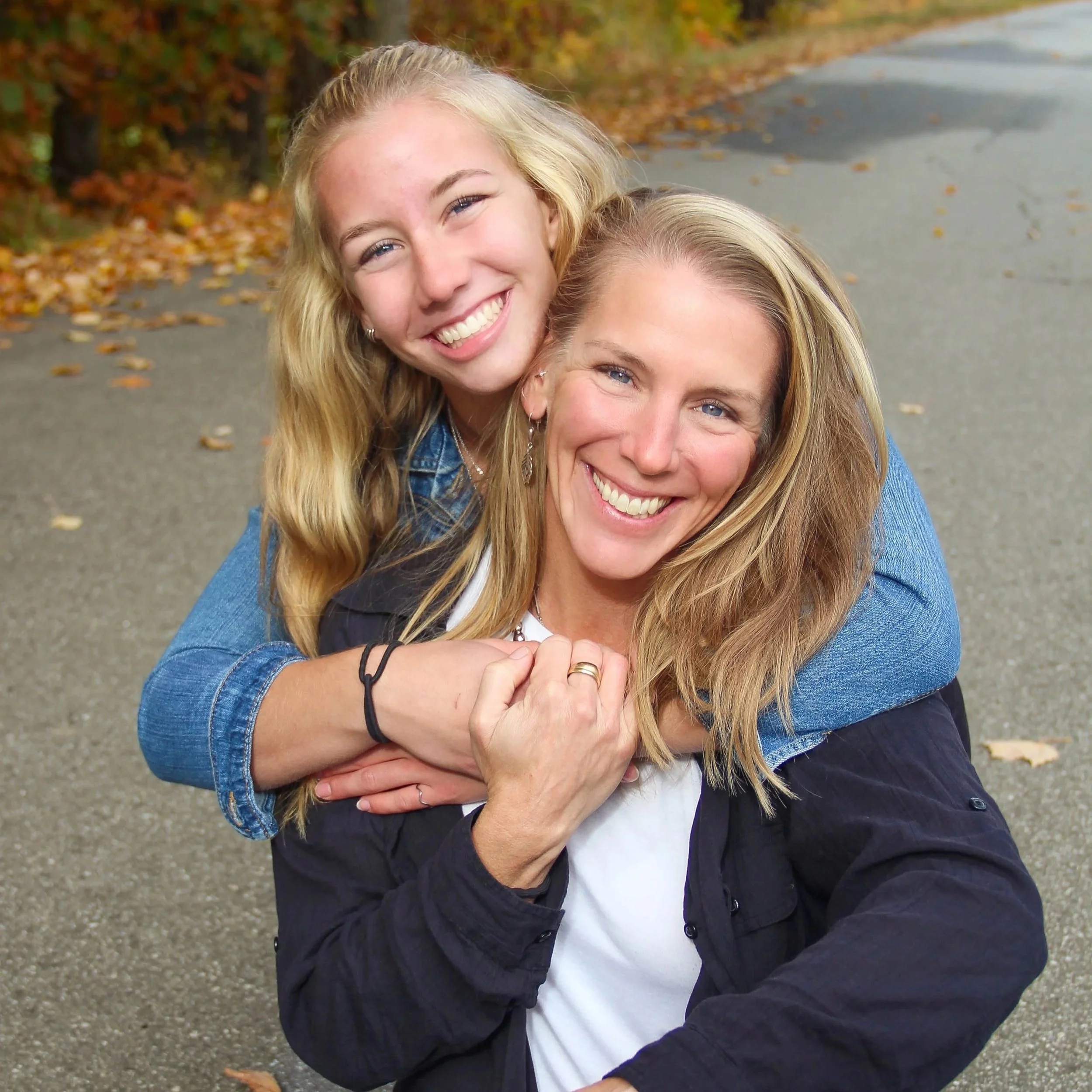A woman with long blonde hair and a woman with long blonde hair are smiling and hugging outdoors, with autumn leaves in the background.