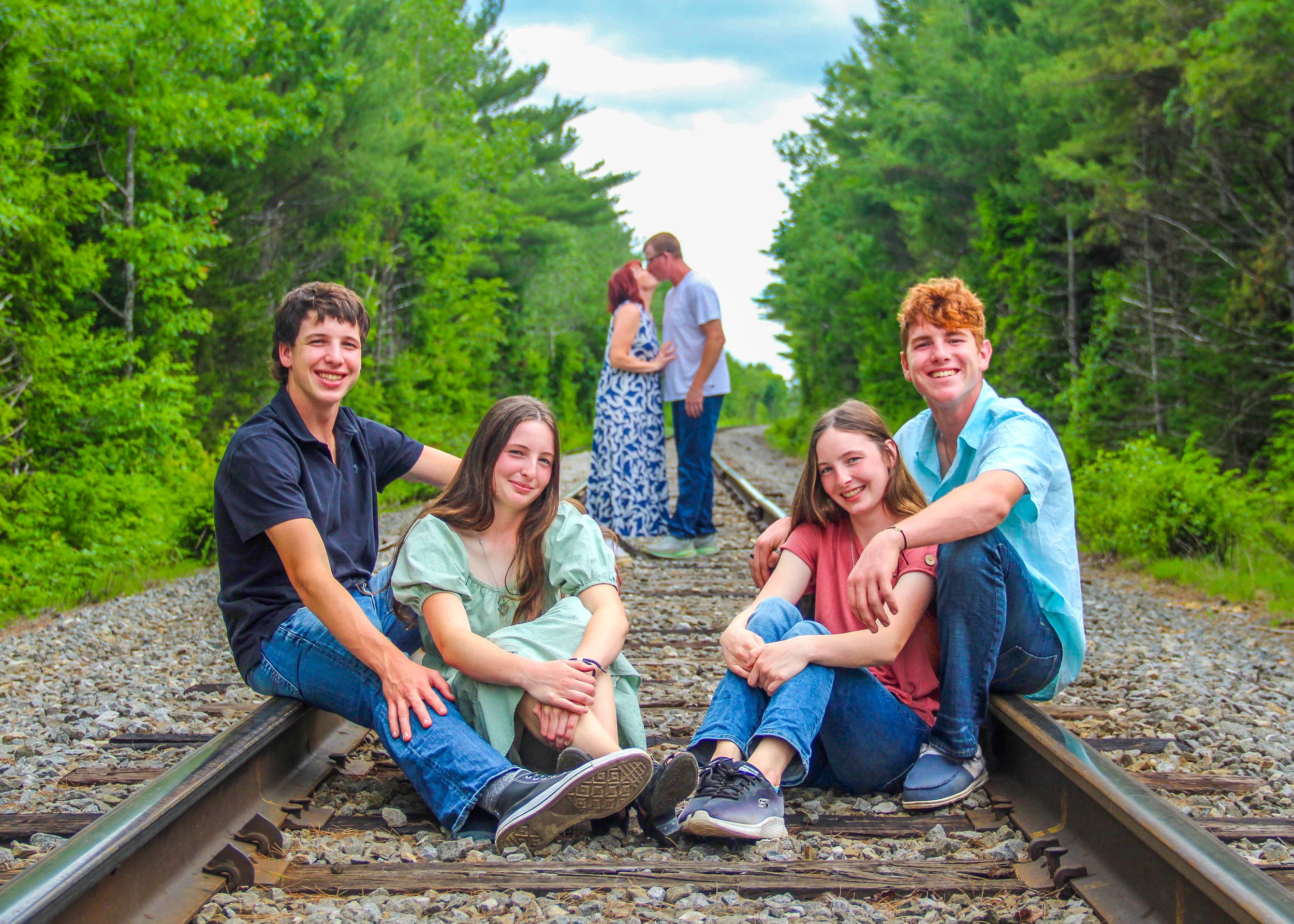 Four teenagers sitting on railroad tracks in a wooded area, smiling at the camera, with a young couple kissing in the distance.