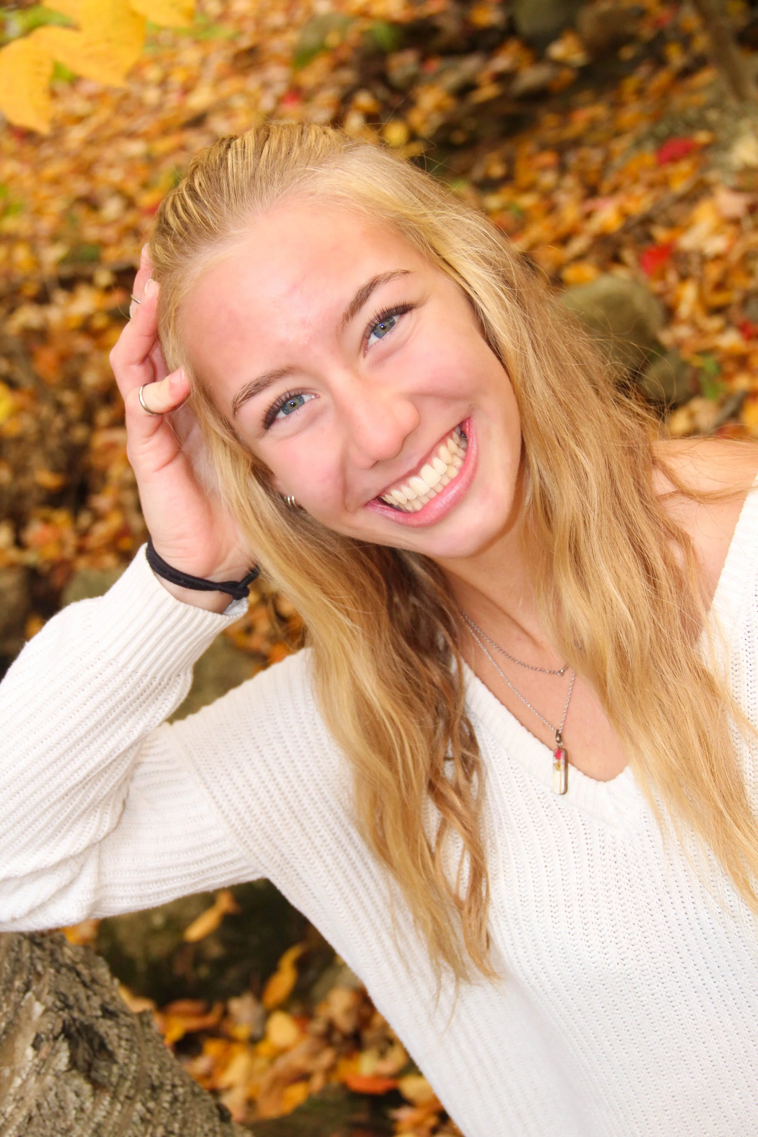 A young woman with long, wavy blonde hair smiling and looking up, wearing a white sweater and a necklace, holding her head with one hand, outdoors surrounded by autumn leaves.
