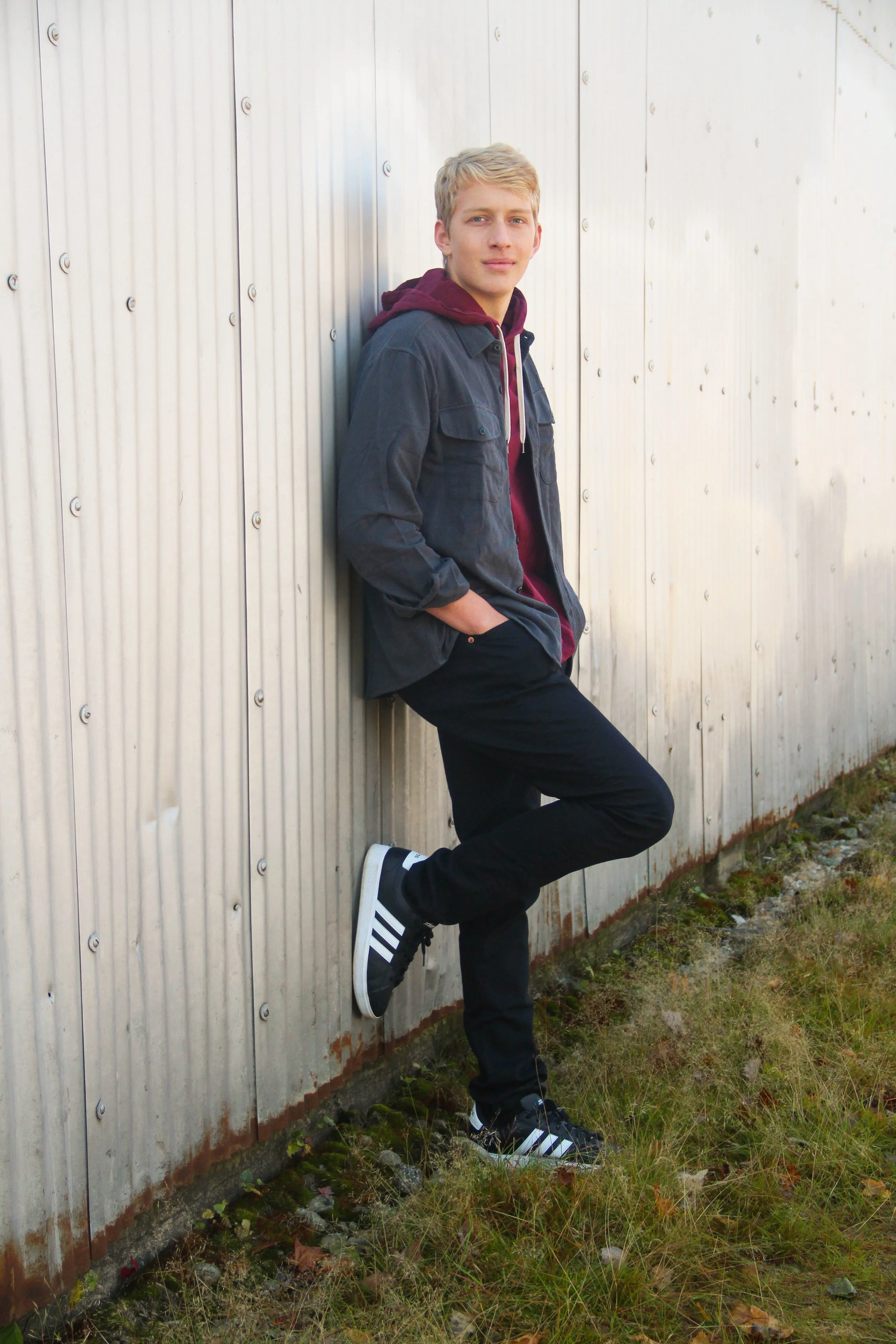 Young man leaning against metal wall, wearing a dark jacket, maroon hoodie, black pants, and Adidas sneakers, with hands in pockets.