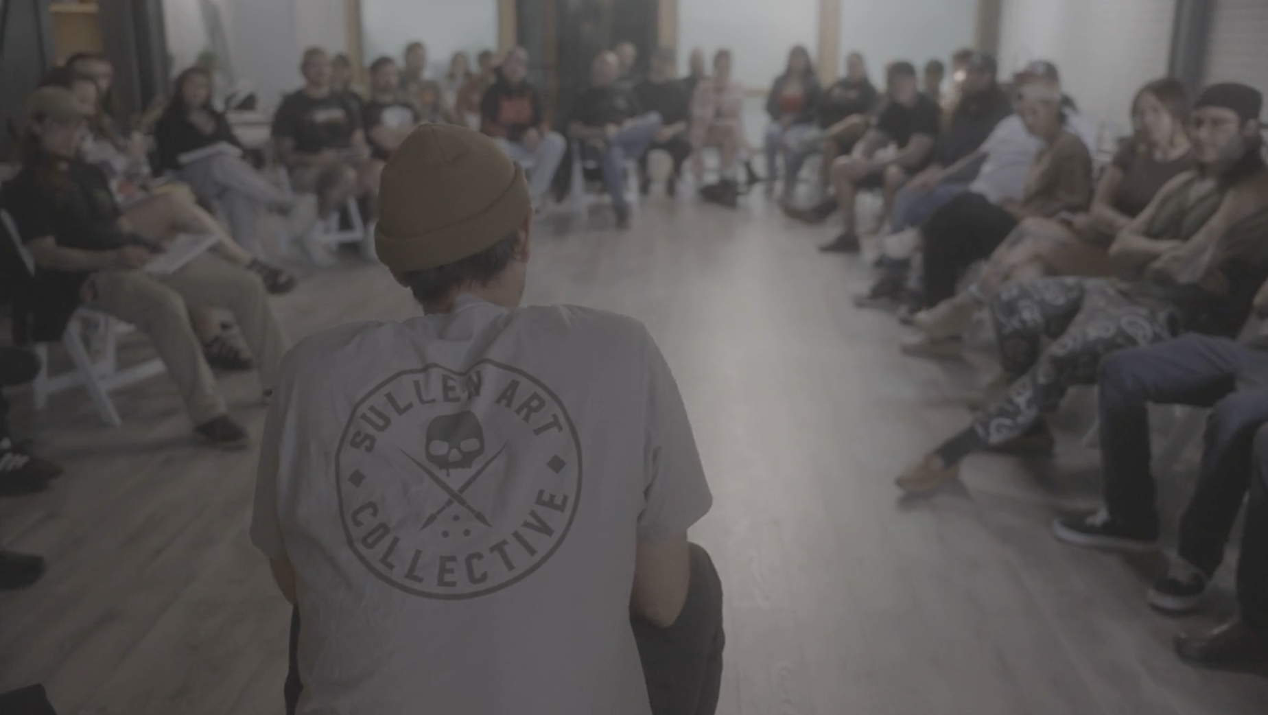 A group of people sitting in a circle in a room, with a person in the foreground wearing a beanie and a shirt with a skull logo and the words 'Sullen Art Collective' on the back.