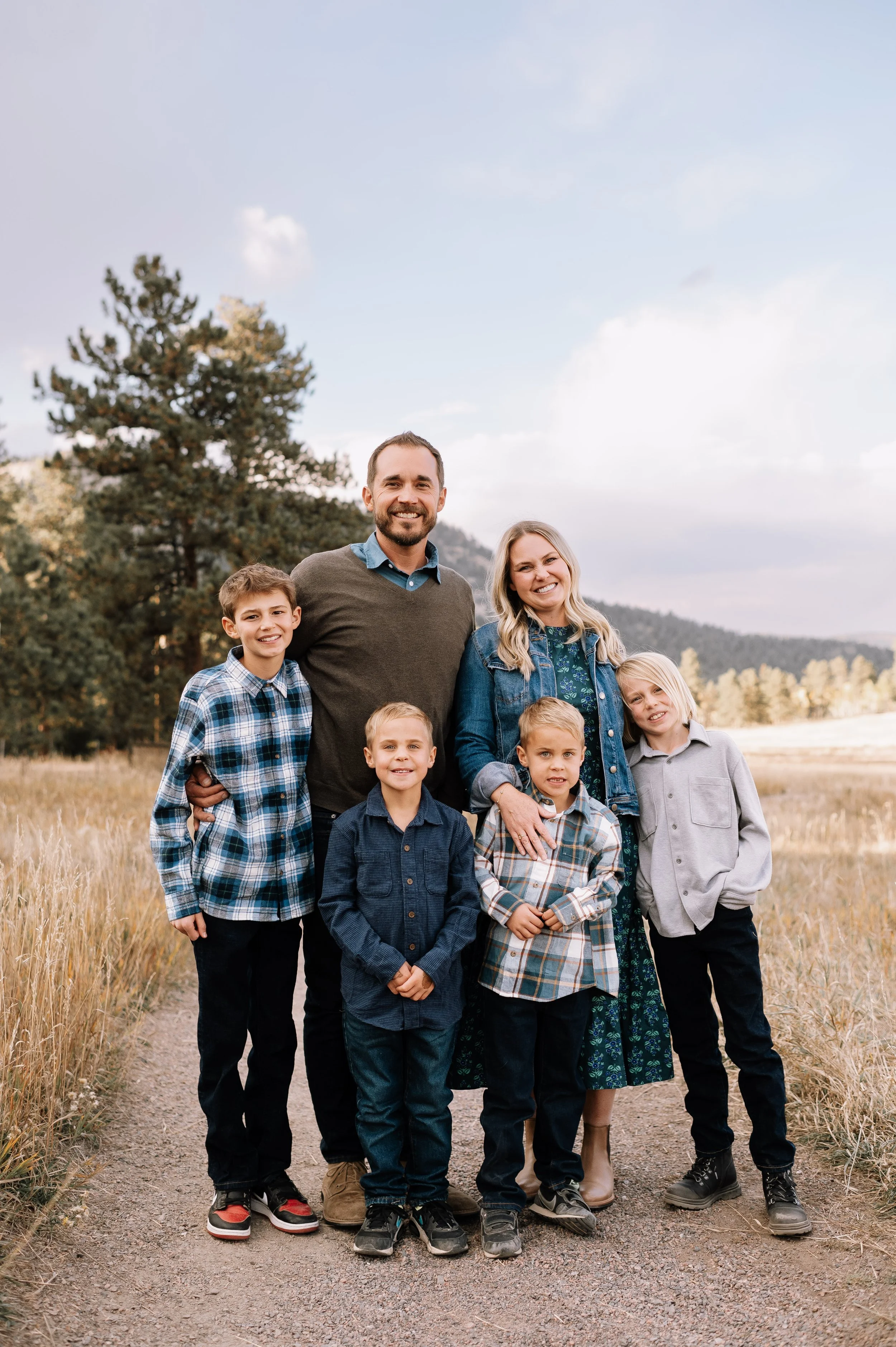 Family standing outdoors on a dirt trail, with trees and hills in the background, smiling at the camera.