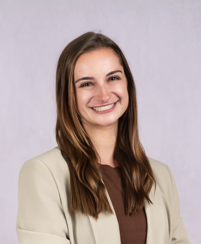 A young woman with long, wavy brown hair and a bright smile, wearing a beige blazer over a brown top, standing against a plain light gray background.