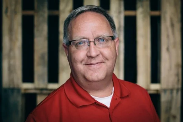 A man wearing glasses and a red polo shirt, smiling in front of a wooden background.