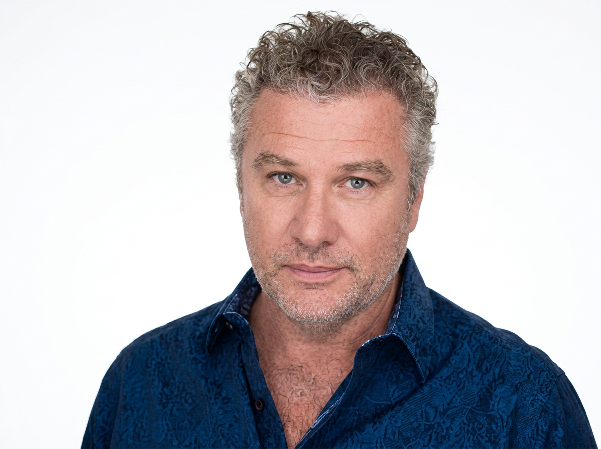 Close-up portrait of a middle-aged man with gray curly hair, blue eyes, and light stubble, wearing a dark blue patterned shirt against a plain white background.