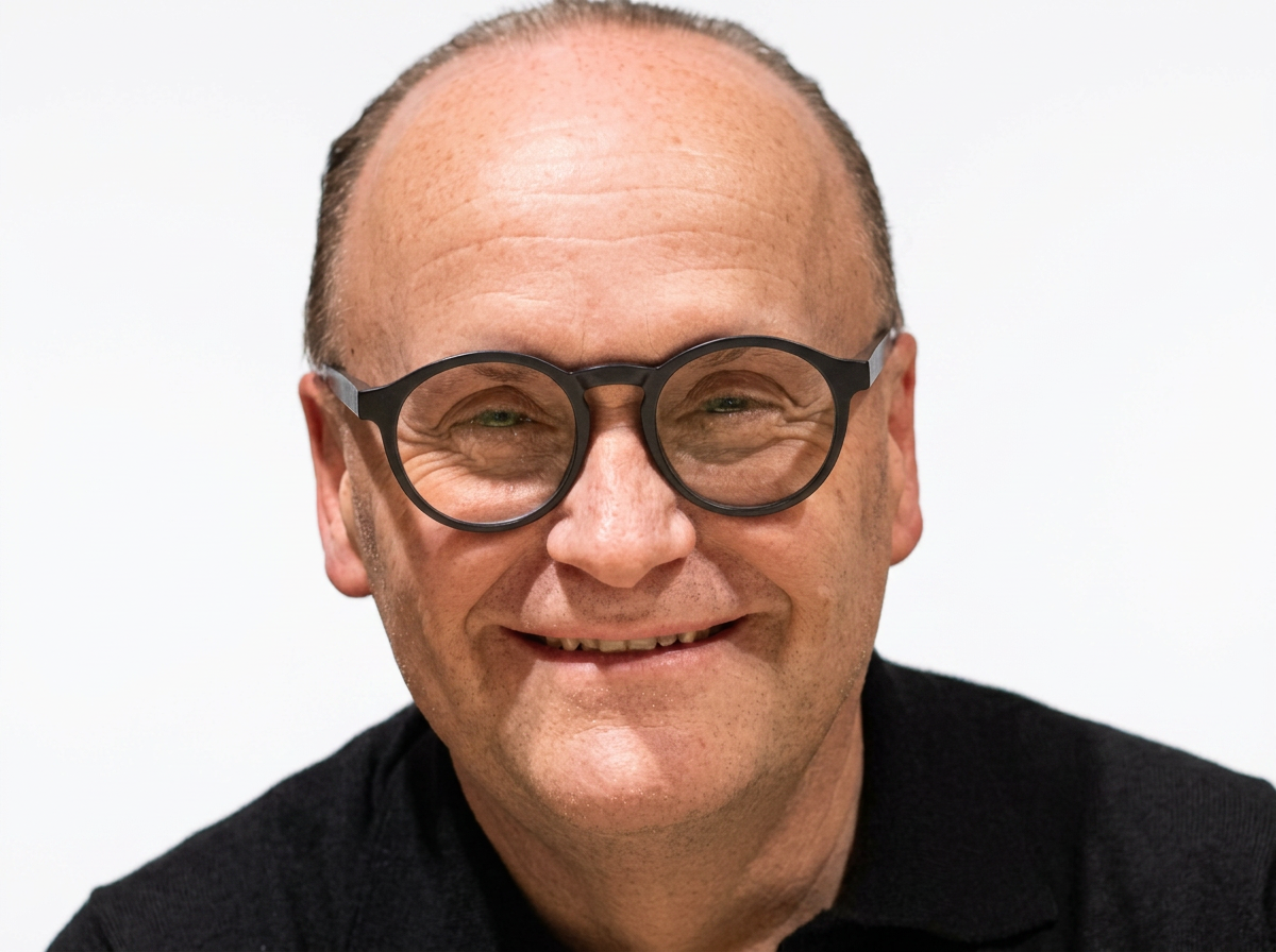 A middle-aged man with light skin, receding hair, and glasses with thick black frames, smiling with his eyes slightly squinted against a plain white background.