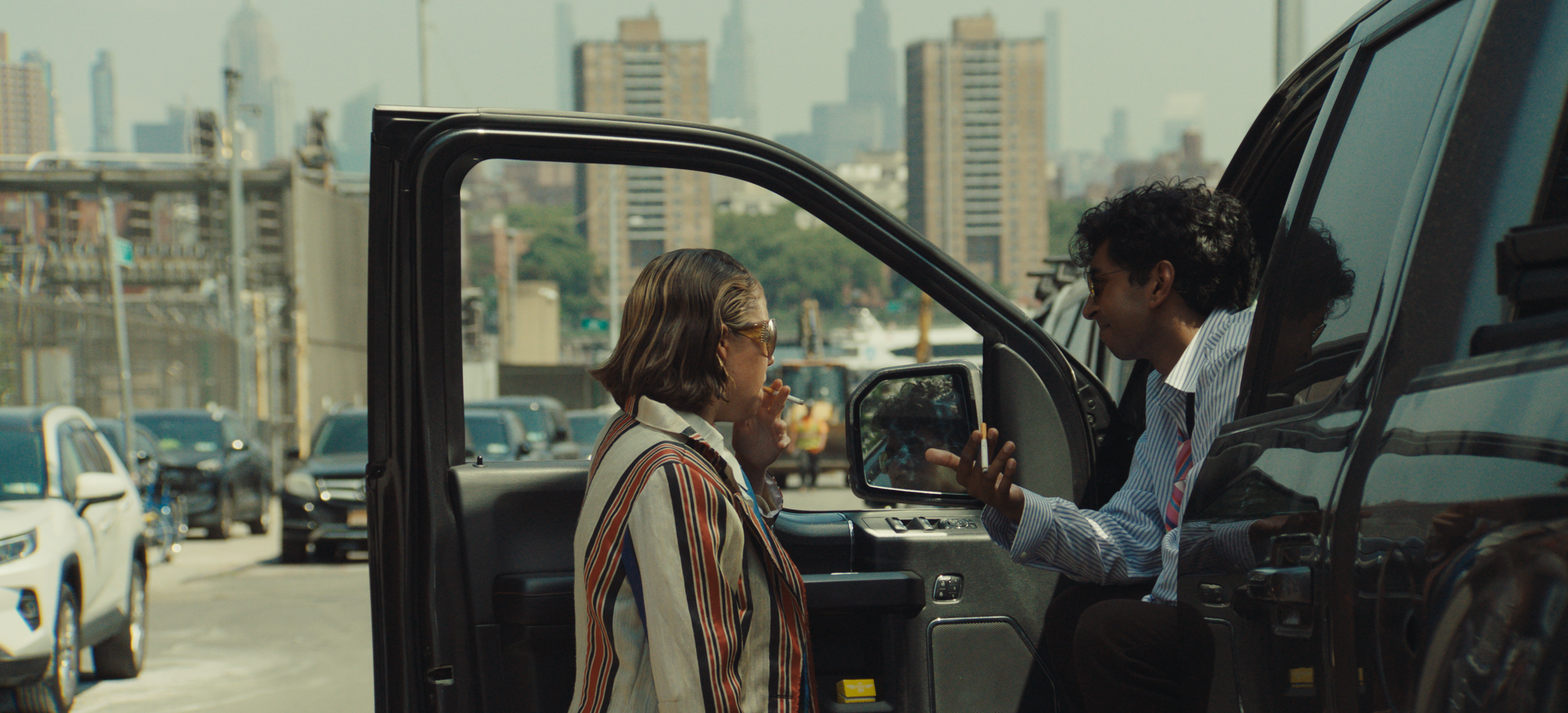 Two people, a woman and a man, are having a conversation near a black vehicle in an urban parking lot with high-rise buildings in the background. The man is showing a phone to the woman, who is listening attentively.