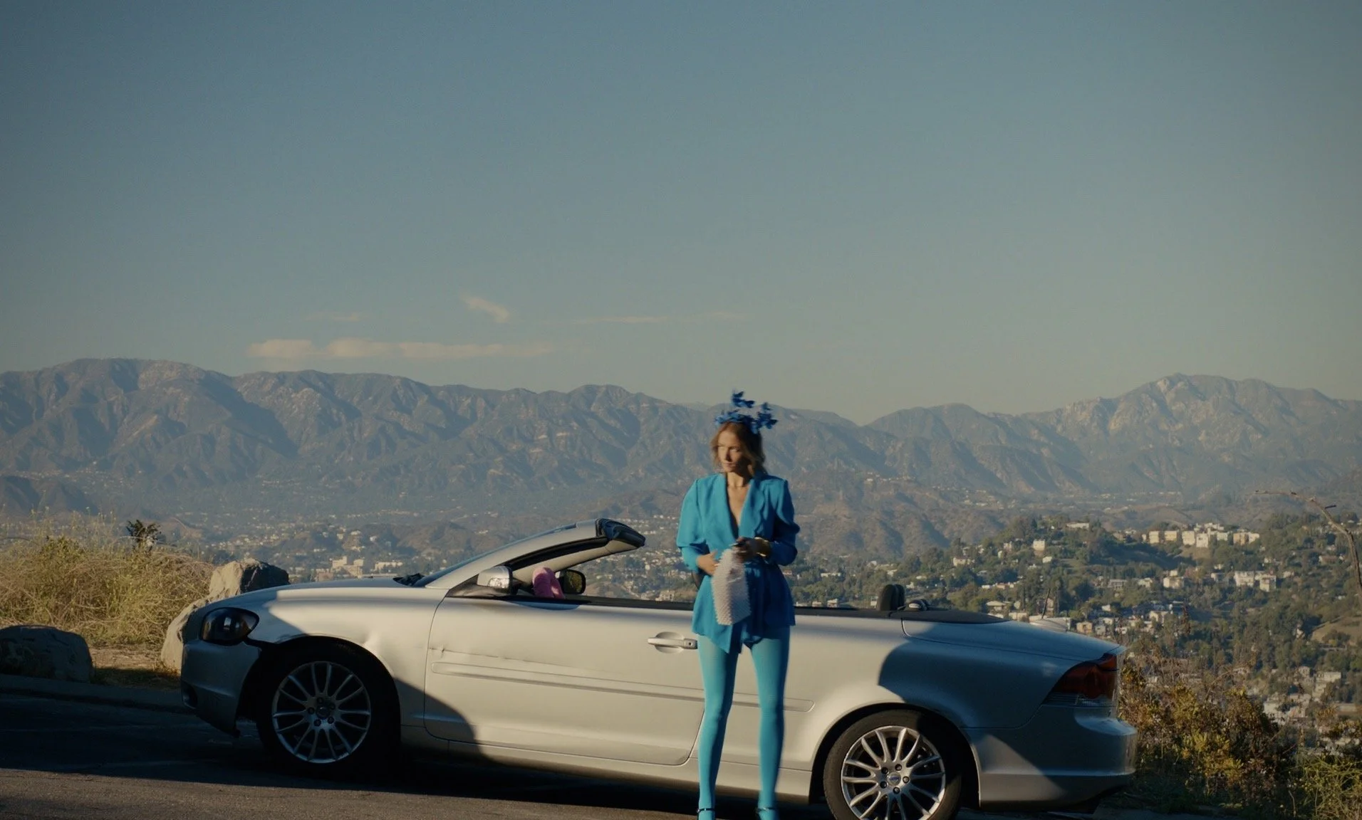 A movie still directed by Libby Hanley of a woman dressed in a blue suit, standing next to a silver convertible car on a scenic mountain overlook, with a city and mountain range in the background.