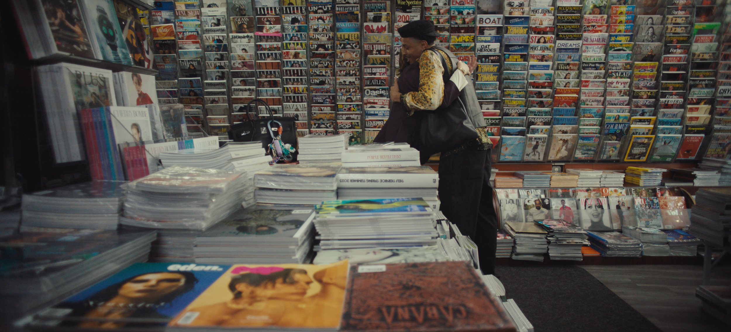 Two people hugging inside a bookstore surrounded by shelves filled with magazines and books, with stacks of magazines and books on tables in the foreground.