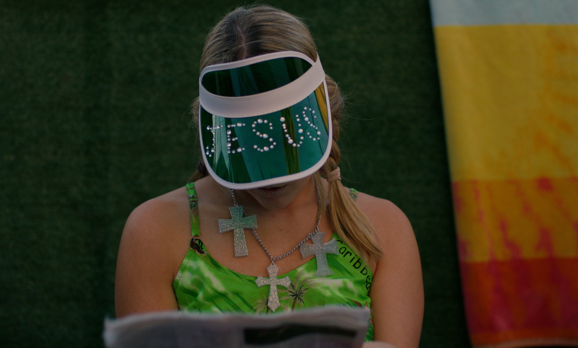 A movie still directed by Libby Hanley of a woman wearing a large visor with the word 'RESIST' written on it, a green tie-dye dress, and multiple cross necklaces, reading a magazine or booklet.