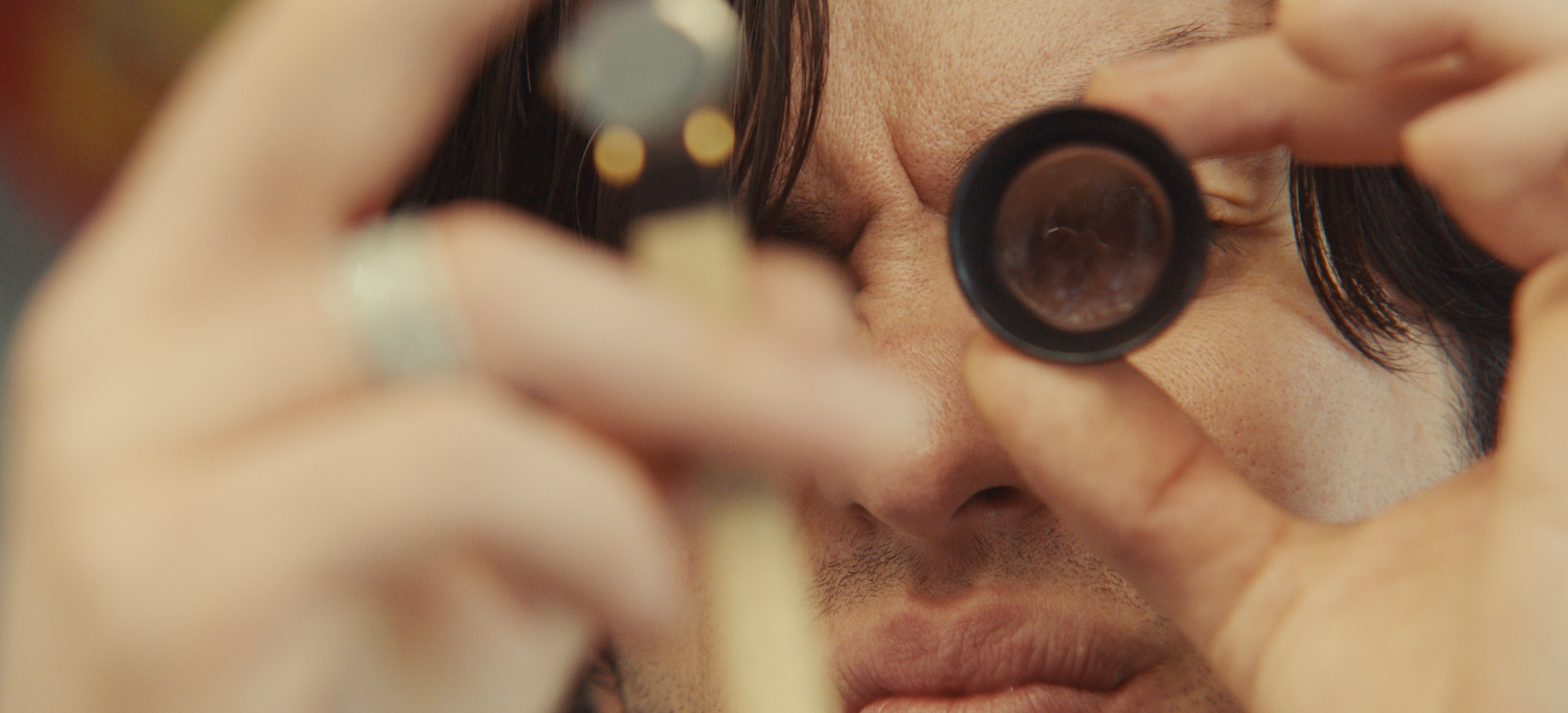 A person is looking through a small, round magnifying lens while holding a small object close to the lens. The person's face is partially visible with dark hair and skin tone.