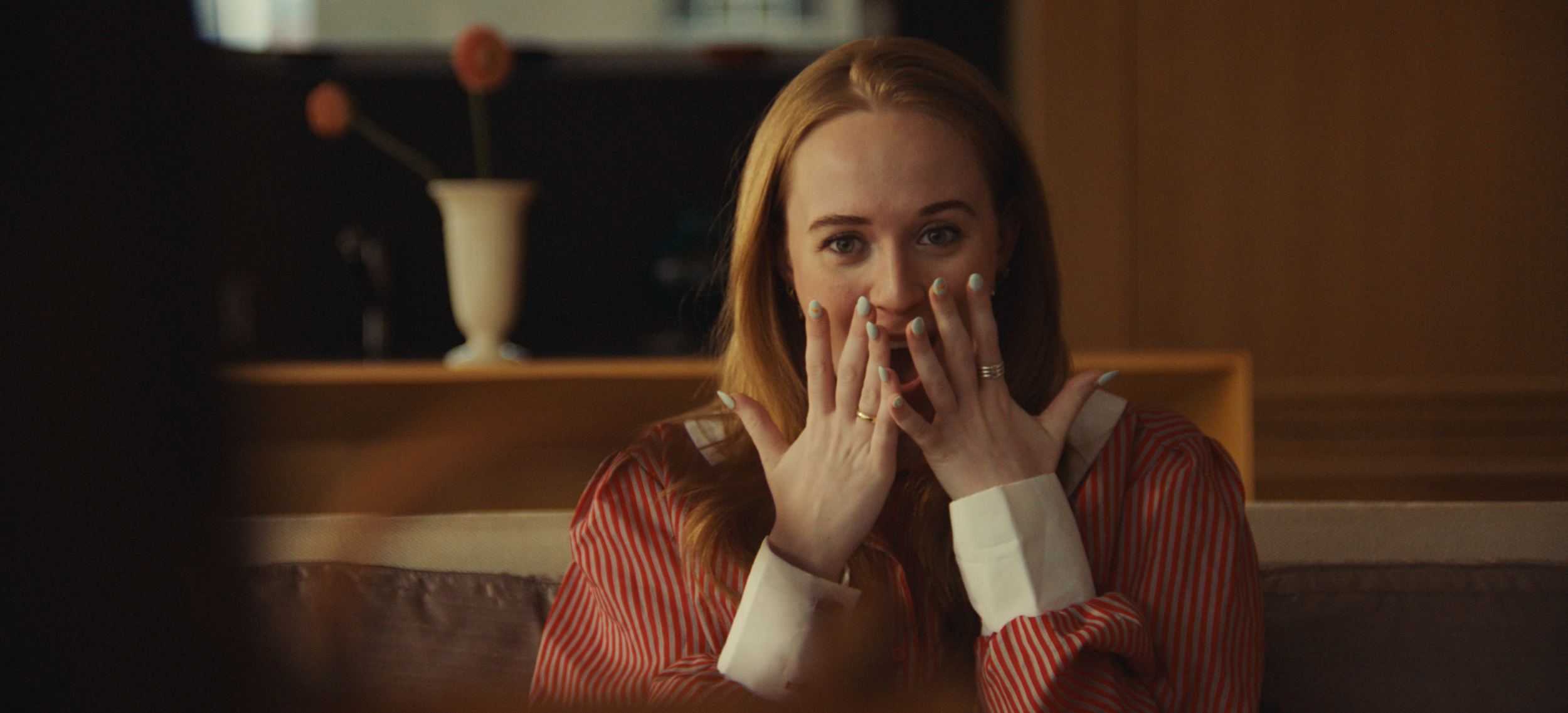 A woman with red hair, wearing a red and white striped shirt, sits at a table with her hands covering her mouth, showing surprised or excited expression.