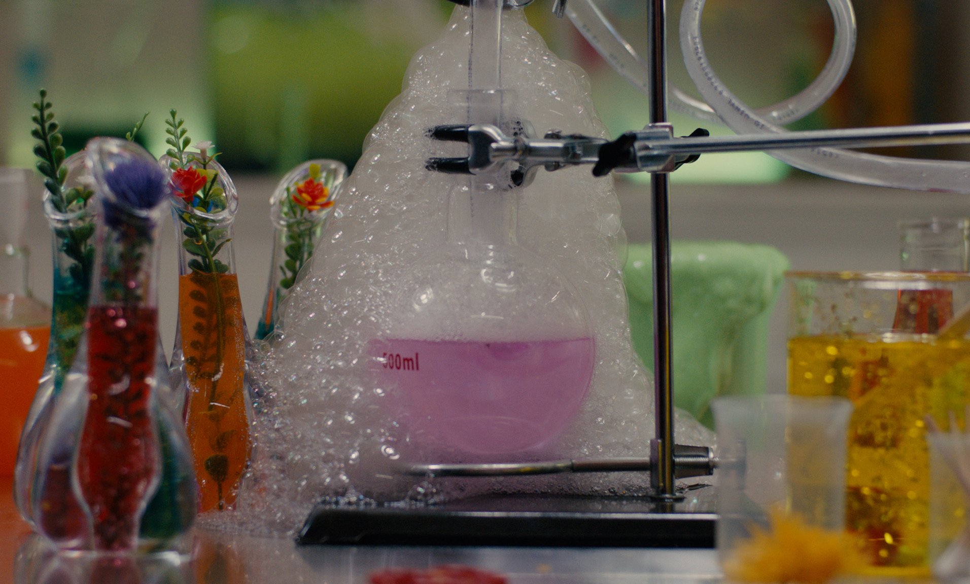 A movie still by Libby Hanley of Science experiment with a boiling flask on a stand surrounded by colorful liquids and decorative plant-like objects.