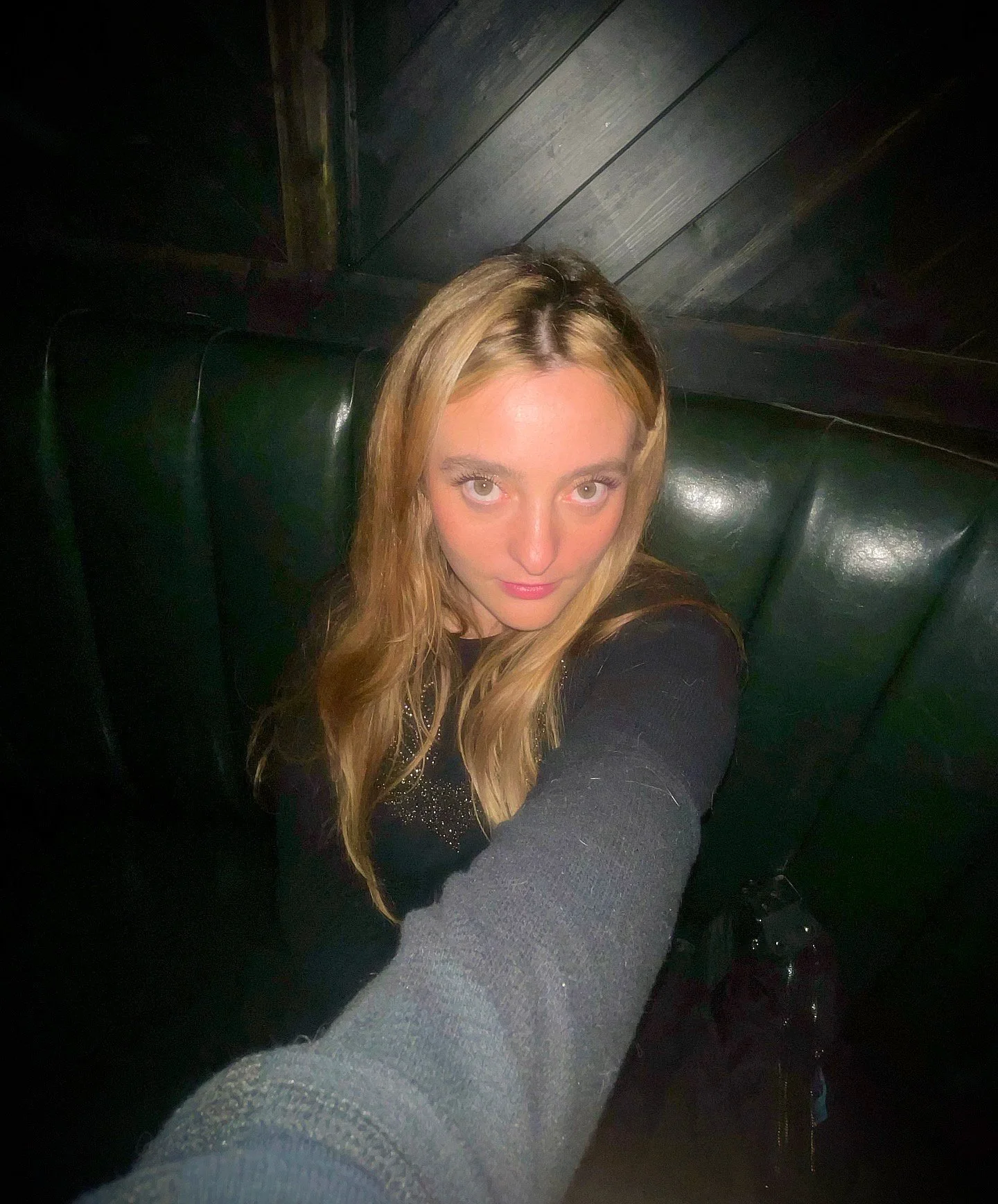 A woman with long blonde hair taking a selfie indoors in front of a dark green booth seat with wood-paneled walls in the background.