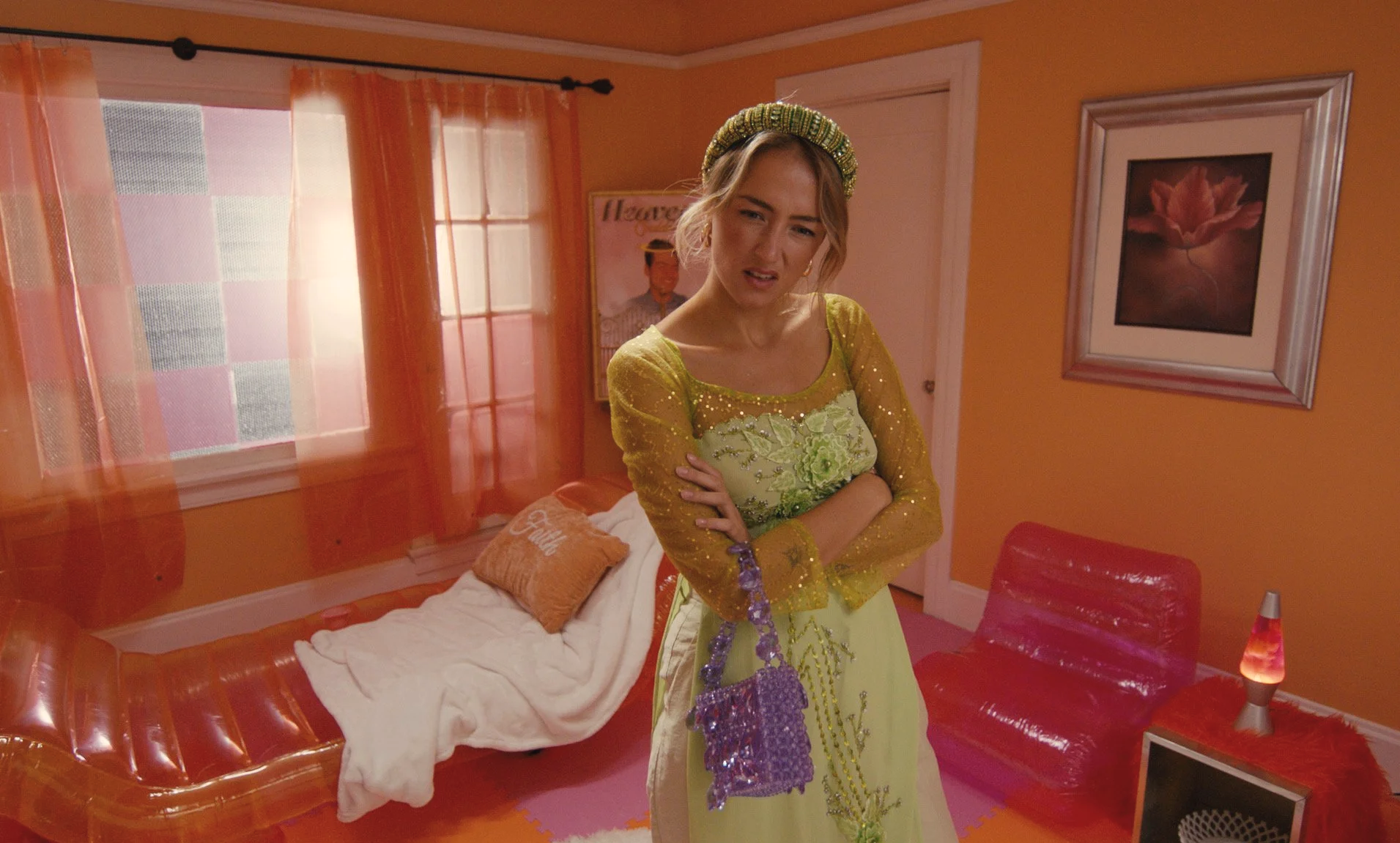 Woman in yellow dress standing with arms crossed in a colorful living room with orange walls, sheer curtains, orange furniture, and framed artwork.