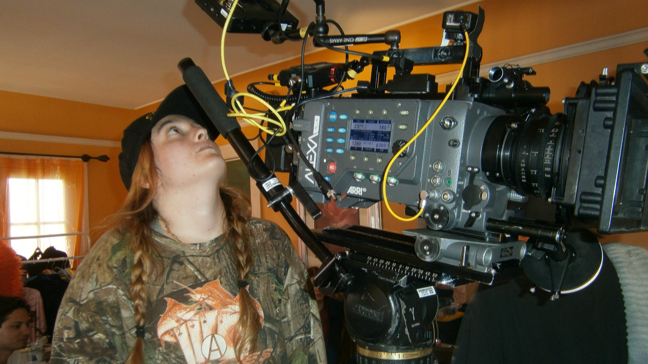 A young woman with long braided hair, wearing camouflage clothing and a beanie, operating a professional film camera indoors.