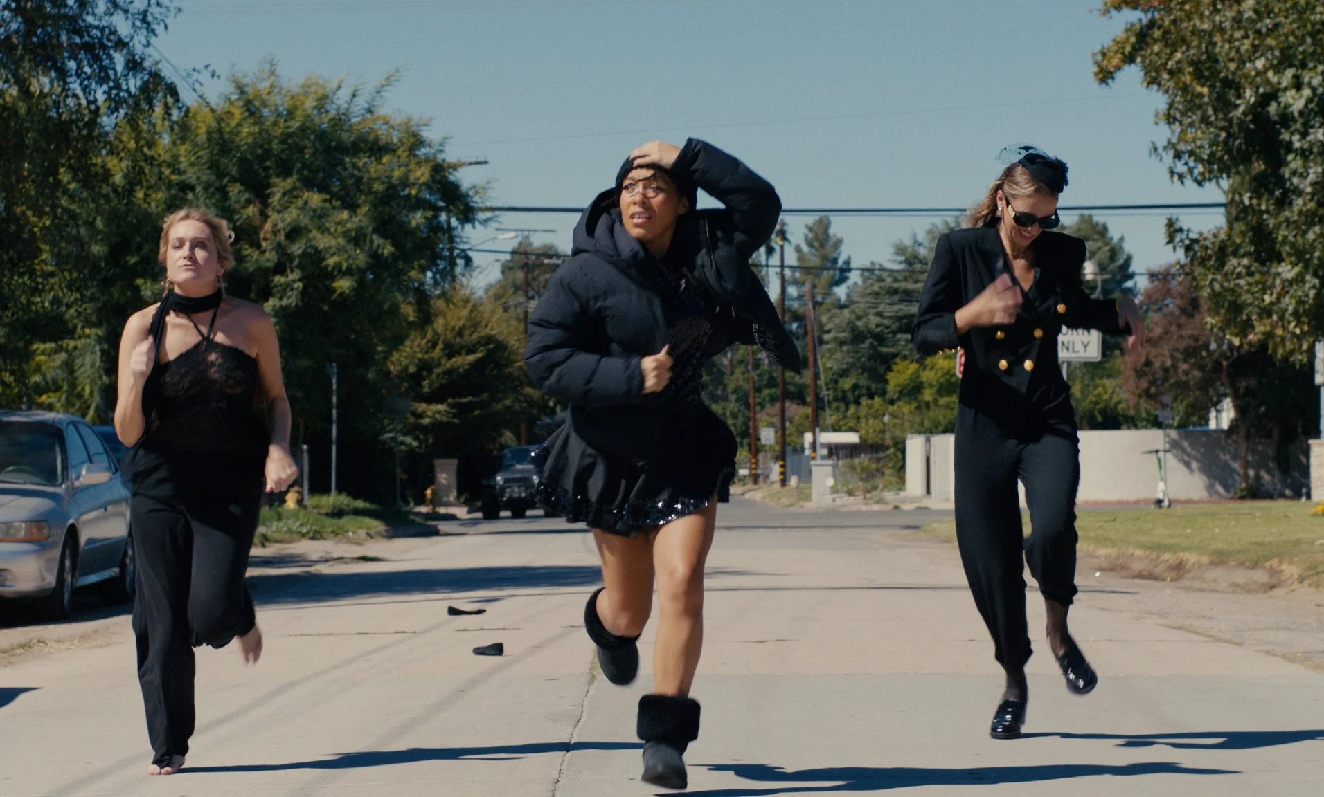 Three women running down the street on a sunny day, dressed in stylish black outfits. The woman in the middle wears a black puffy coat, shorts, and furry boots, her hand on her head. The woman on the right wears sunglasses and a double-breasted black suit. The woman on the left wears a black lace top and loose black pants.