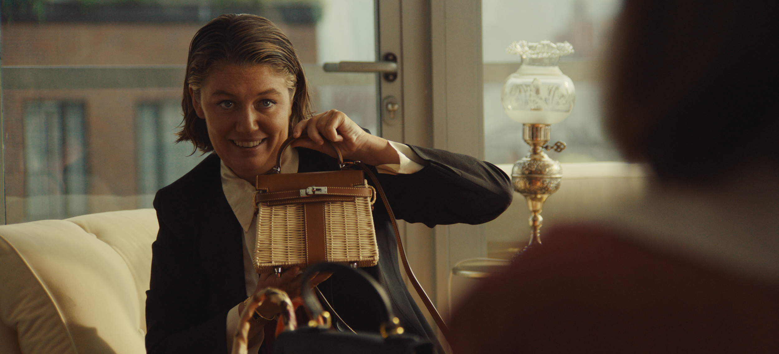 Woman smiling and holding a wicker handbag, seated indoors near a window with a lamp in the background.