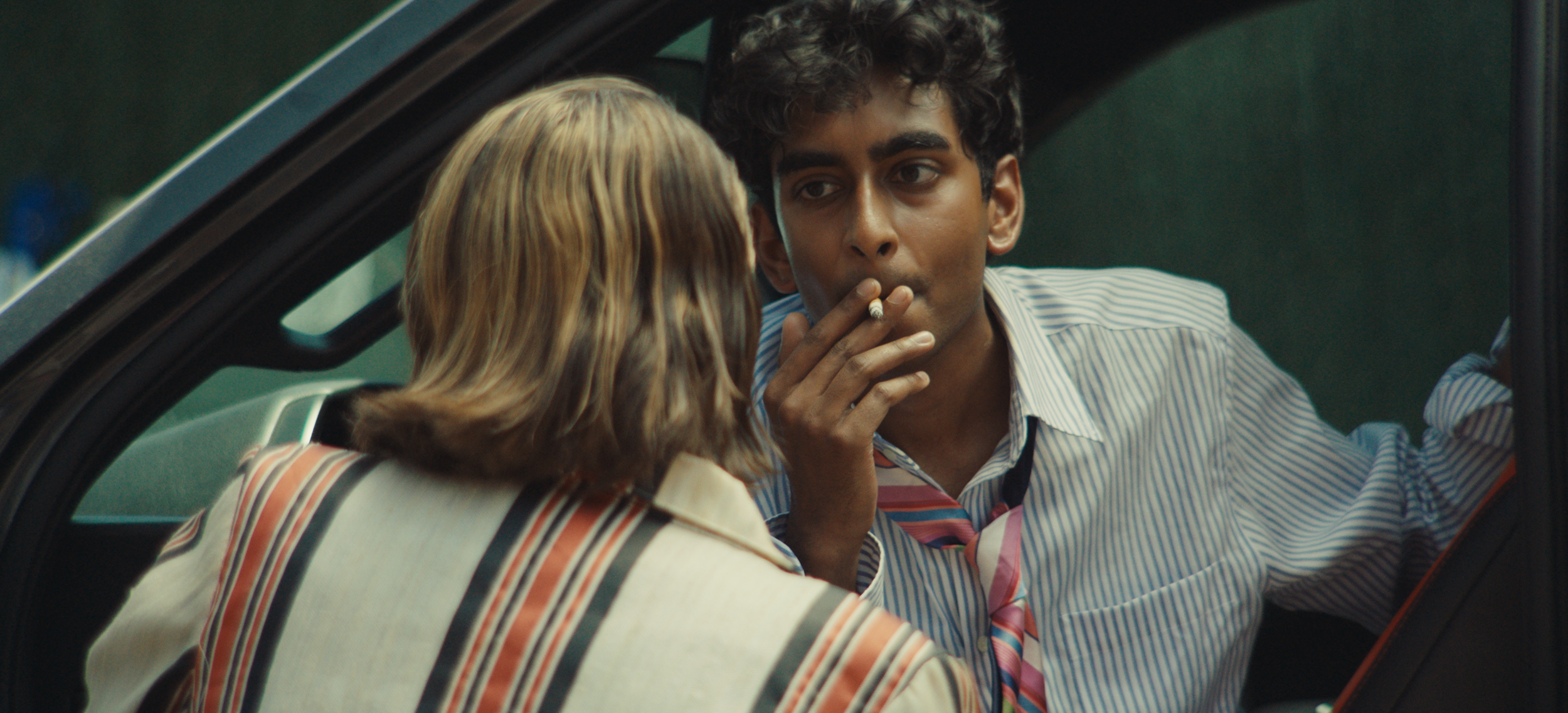 A man with dark curly hair and tan skin smoking a cigarette while talking to a woman with blonde hair in a car.
