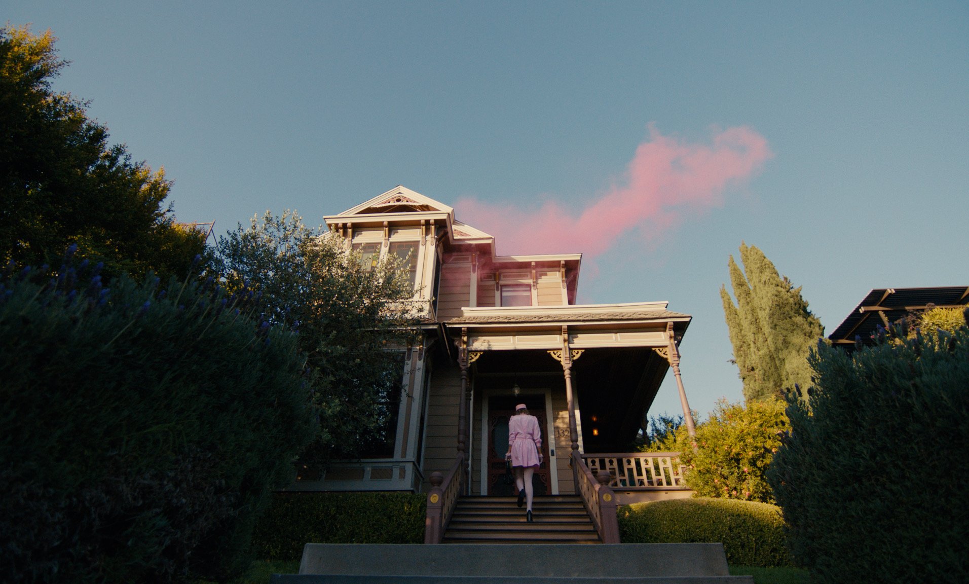 A person dressed in pink walking up the steps to a Victorian house with a green garden and trees around, under a sky with a pink cloud.