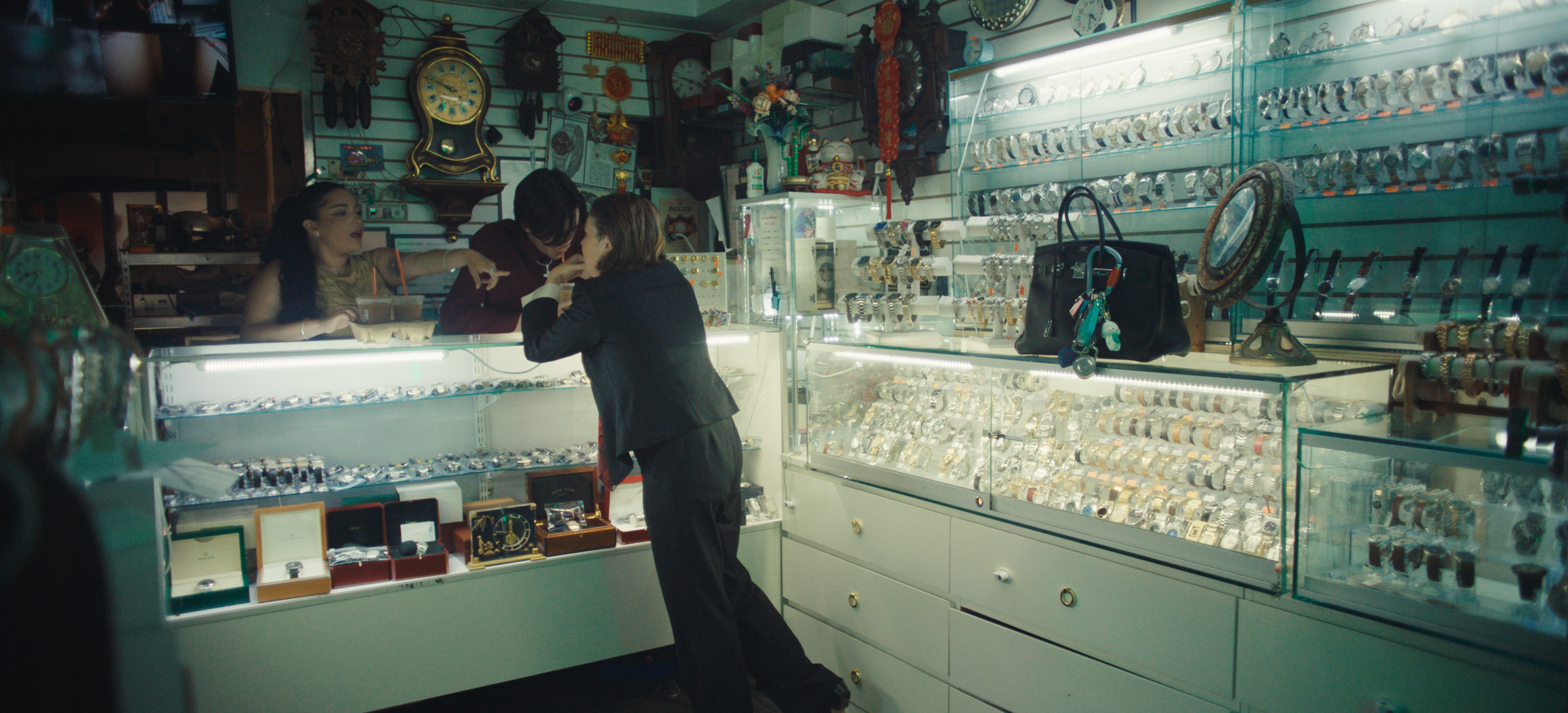 A woman shopping for jewelry at a store counter, engaging with a saleswoman. The store displays various watches, jewelry, and accessories behind glass cases and on shelves.