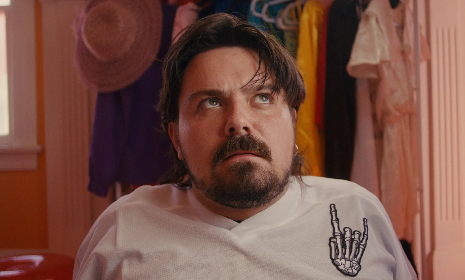 A man with dark hair and a beard making a face with his eyes looking upward. He is wearing a white shirt with a skeleton hand making a rock and roll gesture embroidered on it, and the background shows hanging clothes and a pink wall.