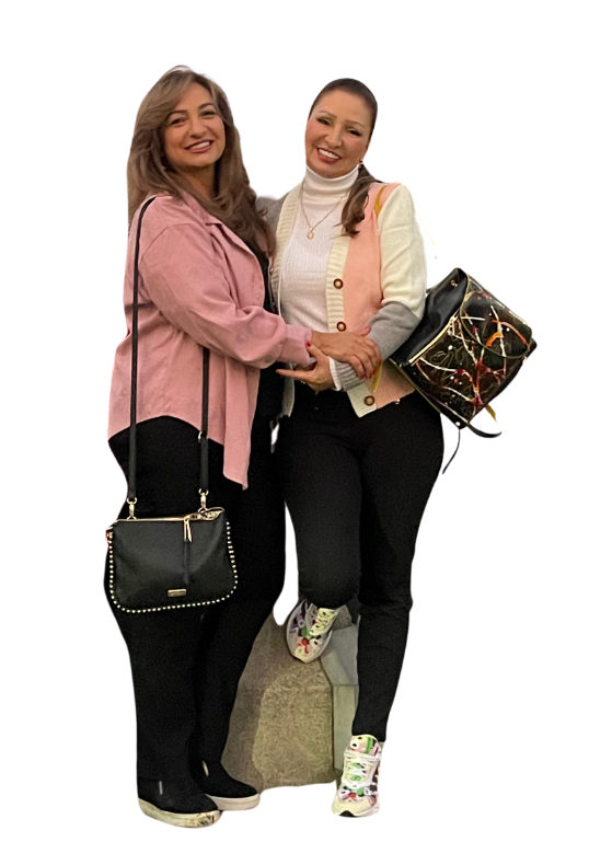 Two women standing together, one wearing a pink jacket, the other wearing a white and pink sweater, both smiling with handbags.