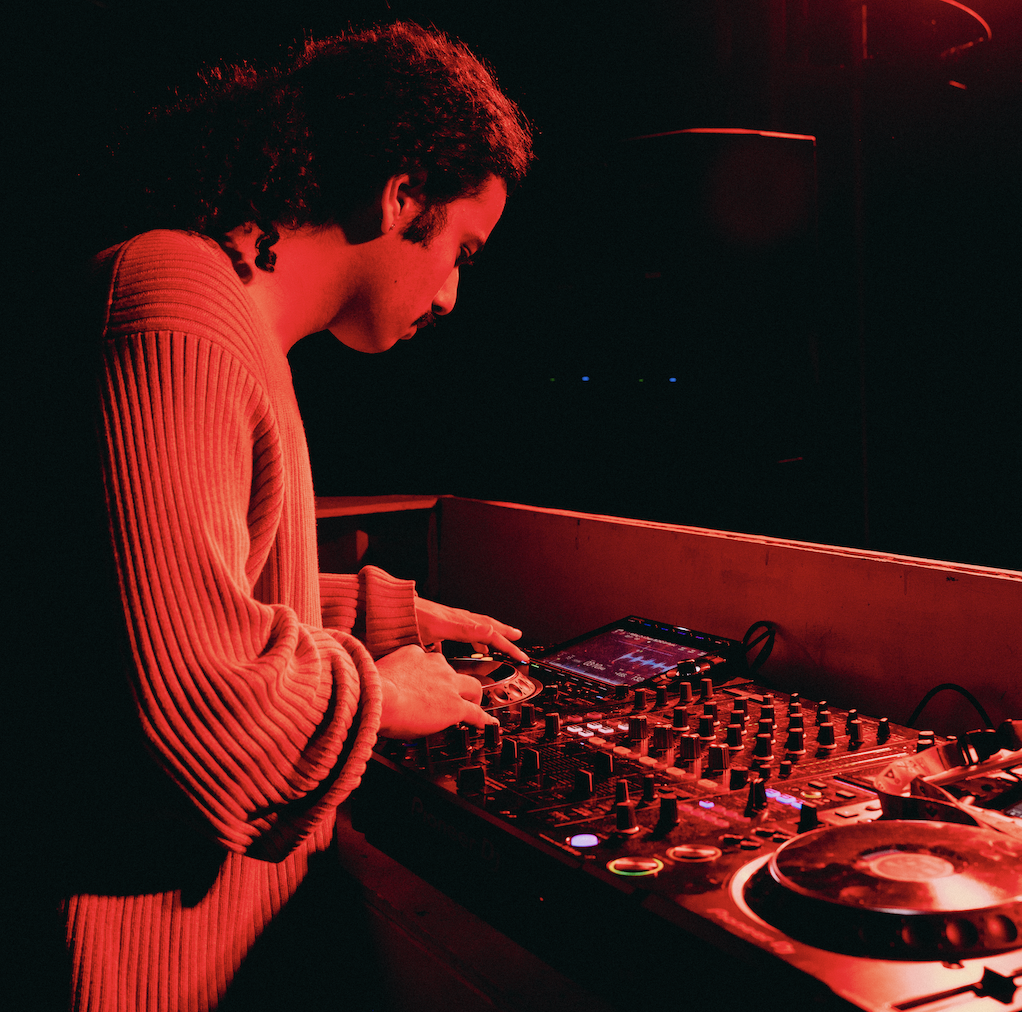 A person with curly hair wearing a red sweater, DJing with a mixing console and a digital screen in a dark, red-lit setting.