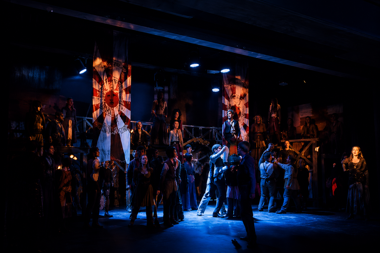 A theatrical stage with many actors in period costumes, illuminated by dramatic stage lighting, with two large banners featuring a stylized sun and face in the background.