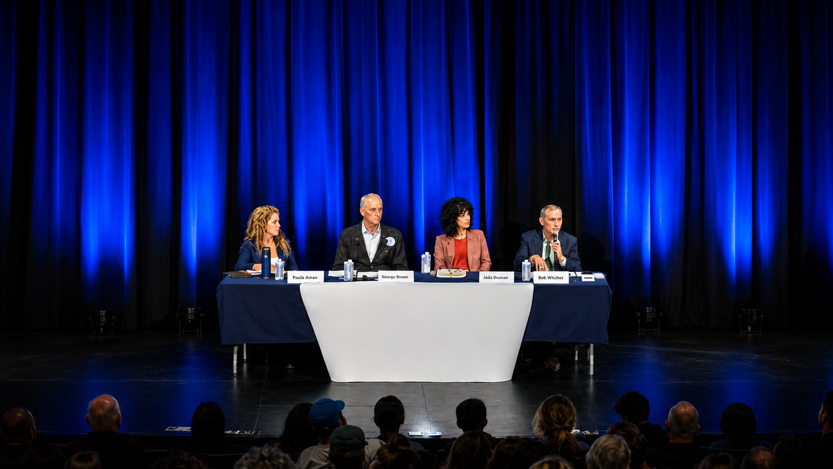 Panel of four people seated at a table on a stage with blue curtains, addressing an audience at a conference or event.
